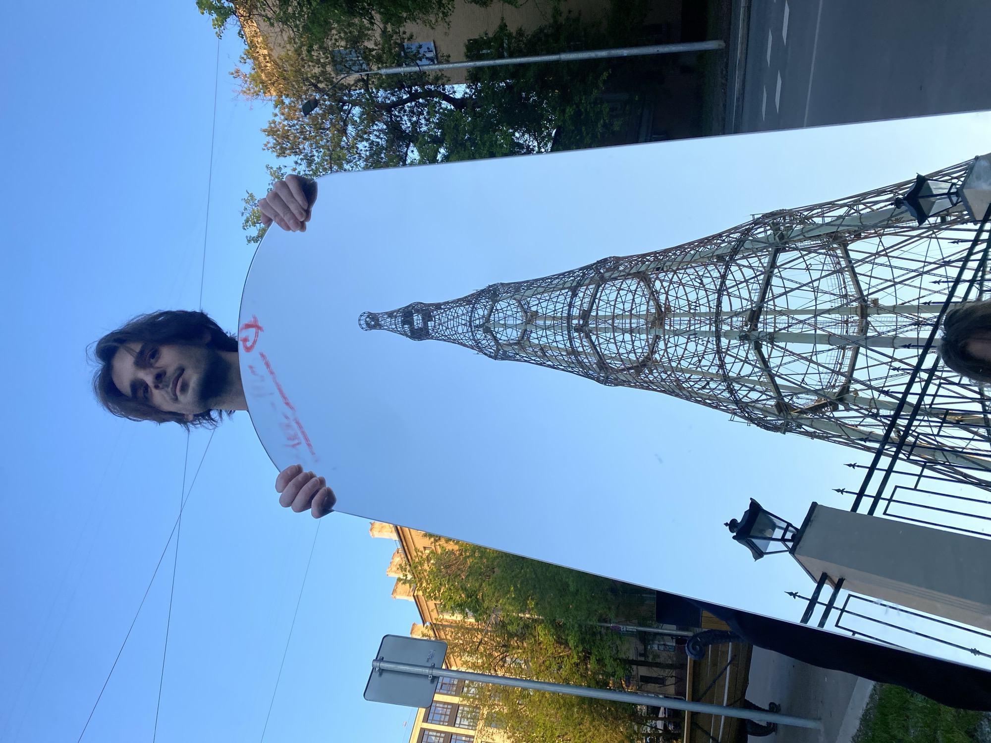 A man holds a rectangular mirror with rounded corners in front of him, reflecting the Shukhov Tower in Moscow, Russia. The tower is a hyperboloid structure made of steel lattice. A street with a road, a pedestrian crosswalk and a fence and a lamppost are visible near the tower. The sky is blue. Trees and a building can be seen in the background. The man has dark hair and a beard. Some indistinct red writing is on the mirror.