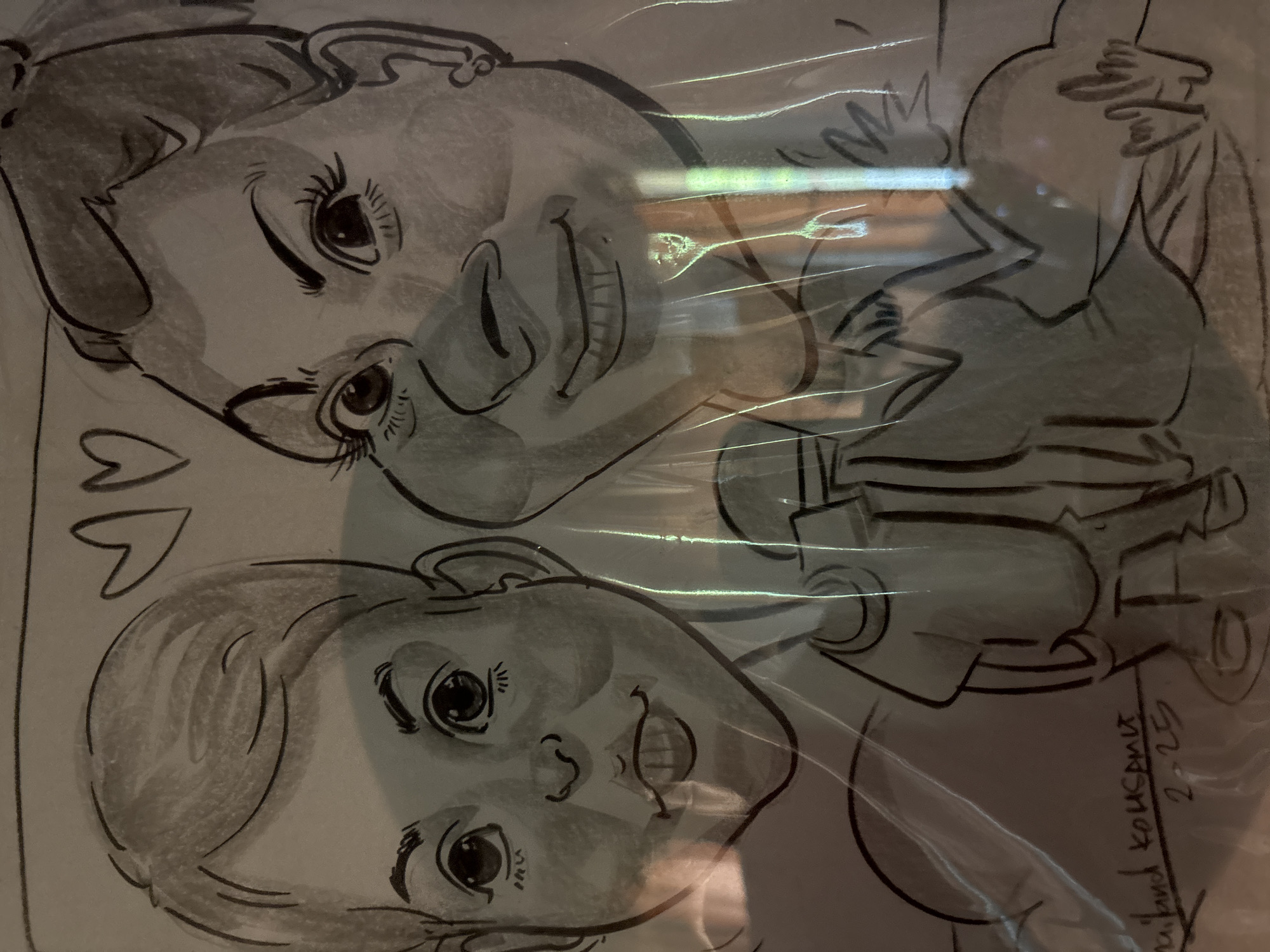 The image depicts a pencil sketch of two children, possibly twins. One child is above the other, and both are facing forward with smiles. There are two heart shapes drawn to the left of the lower child. A figure holding a baby is sketched to the right of the children. There is a signature and date "Roland Kousanik 2-25" in the lower right corner. The artwork appears to be protected by a plastic covering, causing some glare and distortion. The location is บ้านบางรักษ์, Thailand.