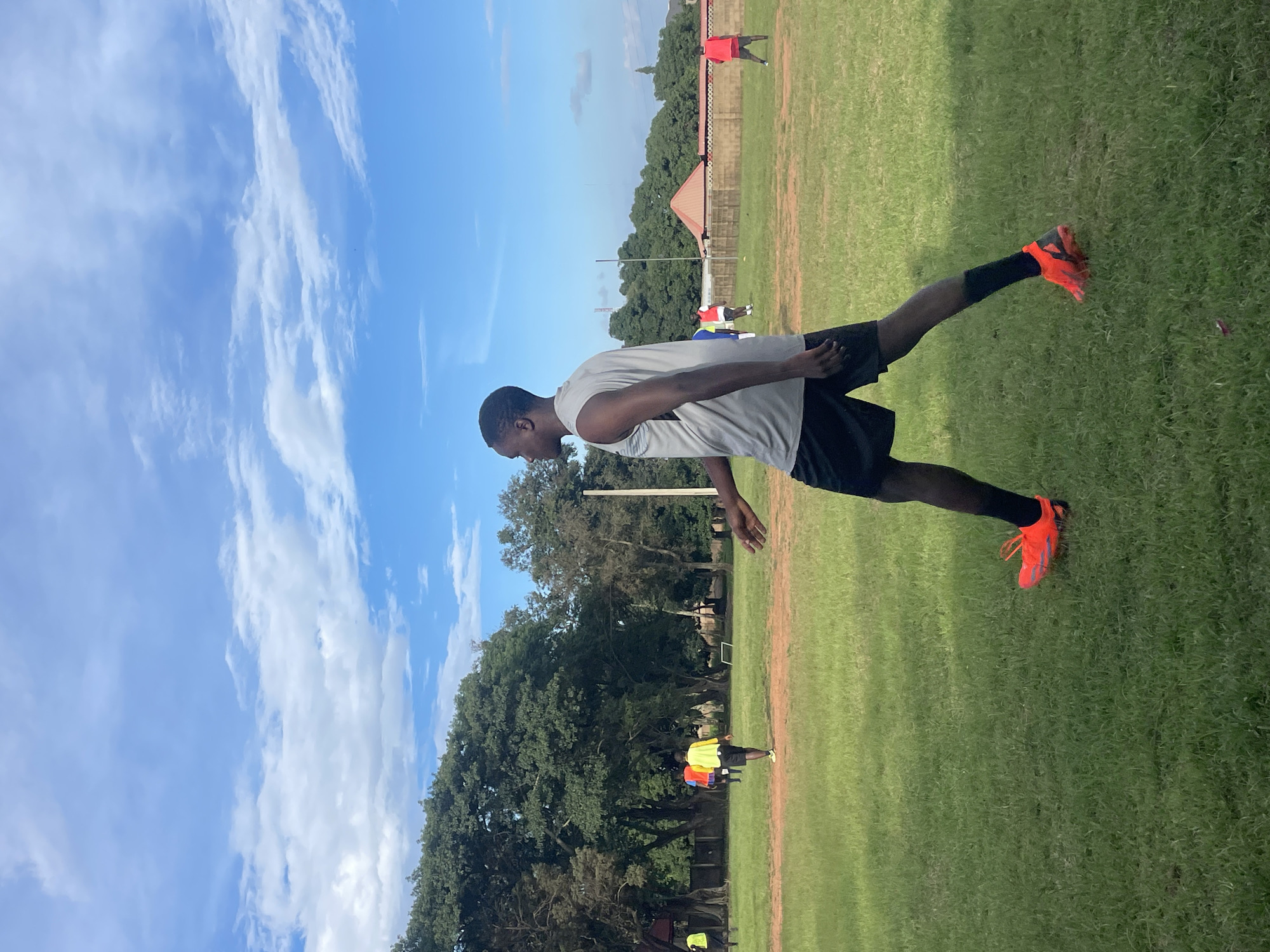 In Jos, Nigeria, on a grassy field under a partly cloudy blue sky, a young Black man is captured in mid-stride, facing left in profile. He is wearing a sleeveless grey athletic top and black shorts, with bright orange athletic shoes and black socks. His right leg is forward, bent at the knee, and his left leg is extended backward. In the background, several other individuals are visible on the field, some wearing brightly colored shirts, suggesting a sporting activity or practice. Lush green trees form a dense backdrop to the scene, with a low wall and buildings further in the distance. The overall impression is of an outdoor recreational setting during daylight hours.