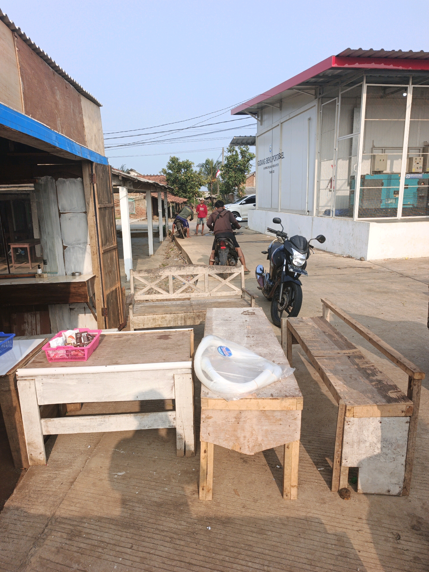 In an outdoor scene in an Indonesian city, a street-level view captures a mix of structures and activity. On the left, a rustic wooden stall with a blue trim is partially visible, displaying a pink basket filled with snacks on its counter. Adjacent to it, a white wooden table with several legs and a clear plastic bag containing a white, coiled item sits on top. Further back, a weathered wooden bench with an ornate design is positioned near a concrete walkway.
Dominating the mid-ground is a black motorcycle, parked on the concrete. A person wearing a brown shirt is seen riding away on another motorcycle in the background, with a white car visible further down the street. To the right, a modern white building with a red roof stands, identified by text that reads "GUDANG BEKU PORT BEL." The building features large white metal fencing. The ground is paved with concrete, showing linear textures and scattered debris. The overall environment suggests a commercial or industrial area with some modest roadside establishments, bathed in the warm light of what appears to be late afternoon.