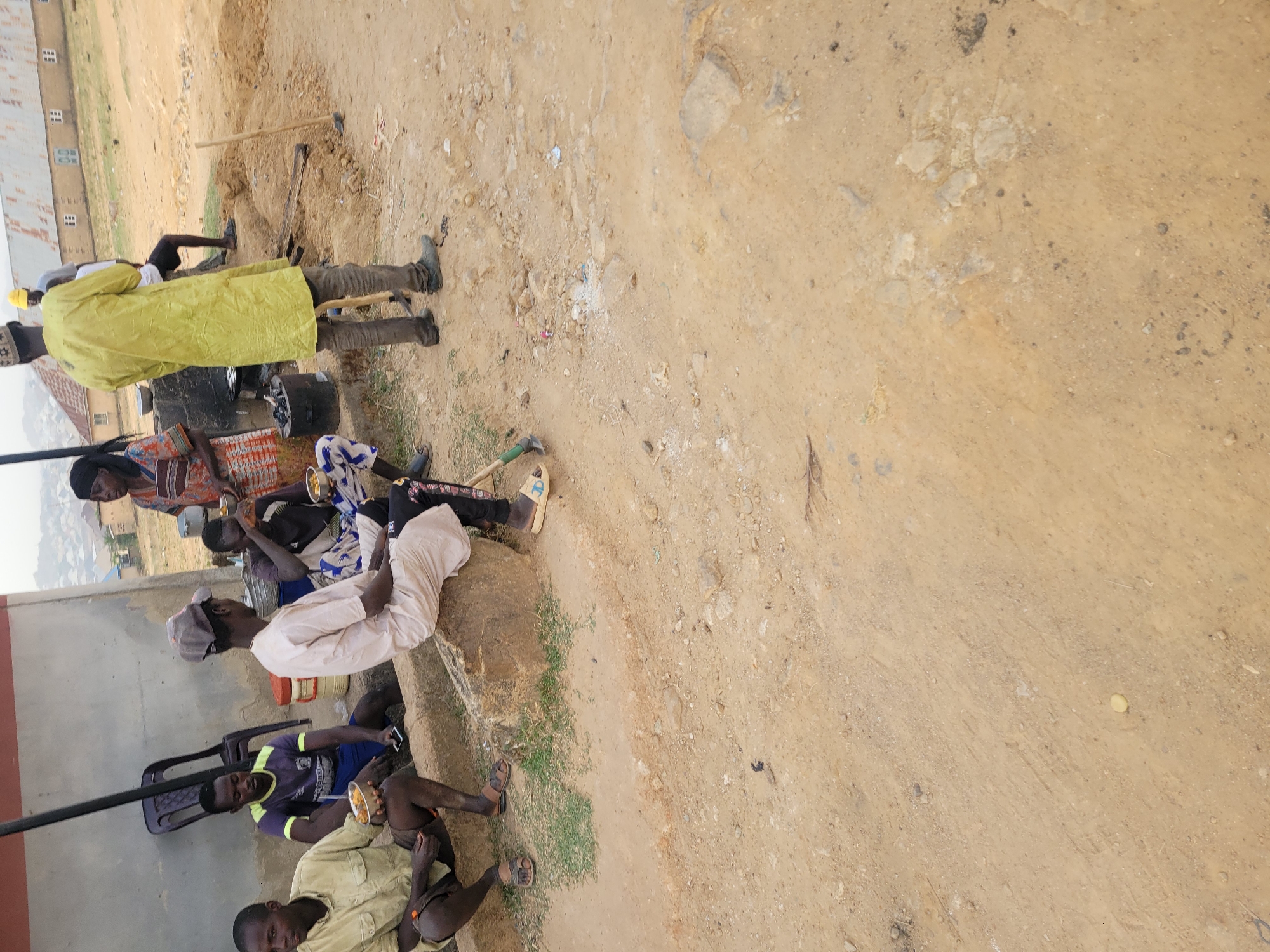 In Barakin Akawo, Nigeria, a group of men are sitting and eating outdoors. Some are seated on chairs, while others are on a raised concrete structure. One man stands near digging equipment. The surrounding ground is a mix of dirt, gravel, and some vegetation. Buildings are visible in the background.