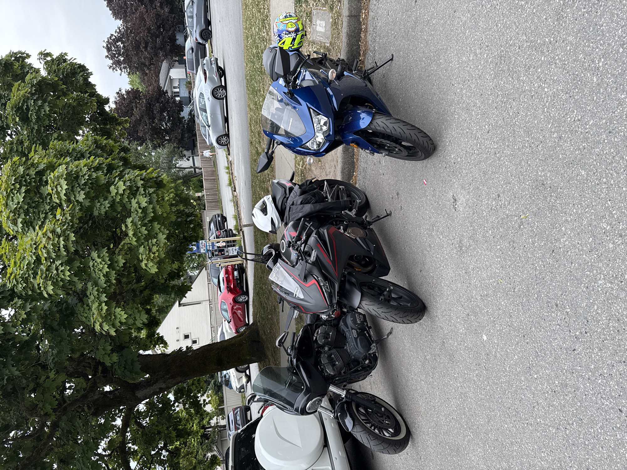 The image shows three motorcycles parked on an asphalt street. The first motorcycle, closest to the camera, is a black motorcycle with a white fairing. The motorcycle in the middle is black with red accents. The motorcycle farthest from the camera is blue and has a helmet on the seat. The street is lined with trees and parked cars. A white building is in the background with a red car visible in front. The location is Vancouver, Canada.