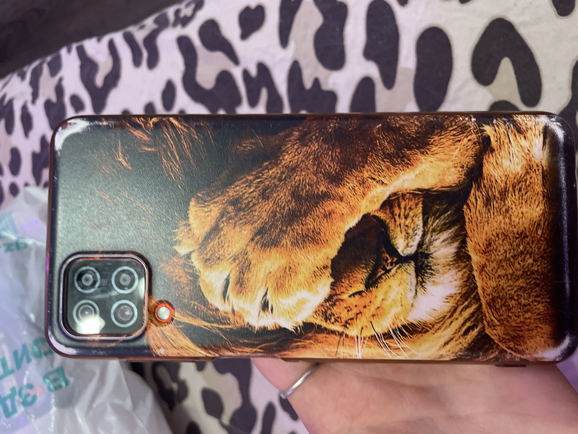 A phone case with a lion's face printed on it is held by a person's hand, which has a silver ring on the index finger. The phone case has a camera module with four lenses at the top. The background is a leopard print pattern. The image is a close-up shot, and the lighting is bright. There are no discernible landmarks or elements that would indicate the specific location of Irinovka, Russia, within the frame.