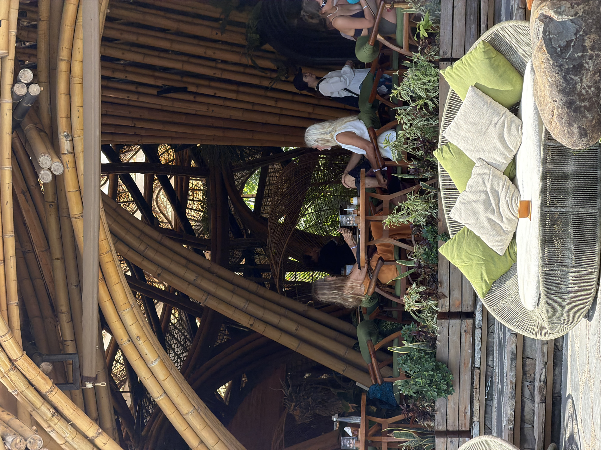 The image depicts an outdoor seating area at a restaurant or cafe in Kediri, Indonesia, characterized by its extensive use of bamboo in its architecture. The structure features a complex, curved roof and wall design constructed from numerous bamboo poles of varying thicknesses and lengths, creating an organic and somewhat enclosed space. Natural light filters through the gaps in the bamboo.
In the foreground, a circular outdoor lounge area is visible, furnished with a rope-woven sofa adorned with several light-colored and green throw pillows. A large, textured rock sits to the right of the sofa.
Further back, several tables and chairs are arranged. Two women are seated at one table on the left, engaged in conversation and looking at their phones. Another table is occupied by a man with long blonde hair, also looking at his phone. Behind them, a woman in a white top and a black cap stands near a table, and further to the right, another woman wearing a black top and sunglasses is seated. A variety of plants are arranged in wooden planters along the edge of the seating area. The overall atmosphere suggests a relaxed, tropical dining or social environment.