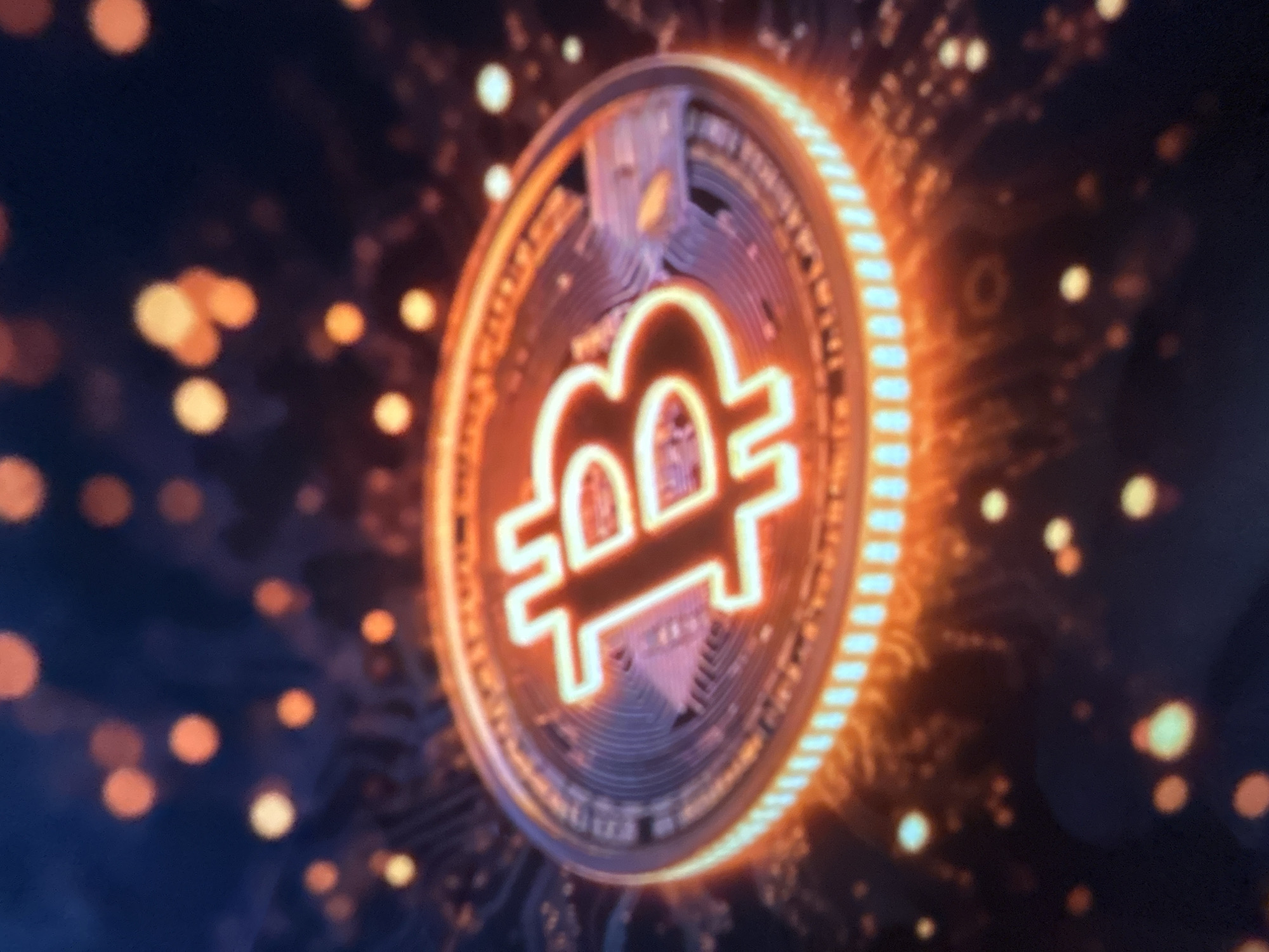 The image displays a close-up, high-angle shot of a stylized, glowing Bitcoin cryptocurrency coin. The coin is depicted in a three-dimensional render, with the iconic "B" symbol prominently featured in the center, outlined in a bright orange neon light. The edge of the coin is also illuminated with a similar orange glow, and the surface of the coin shows intricate, metallic detailing. The background is dark and out of focus, with scattered, blurred points of light in shades of orange and yellow, suggesting a digital or abstract environment. There is no visible connection to the city of Gelendzhik, Russia, or any discernible elements of an urban setting. The image is purely a visual representation of digital currency.