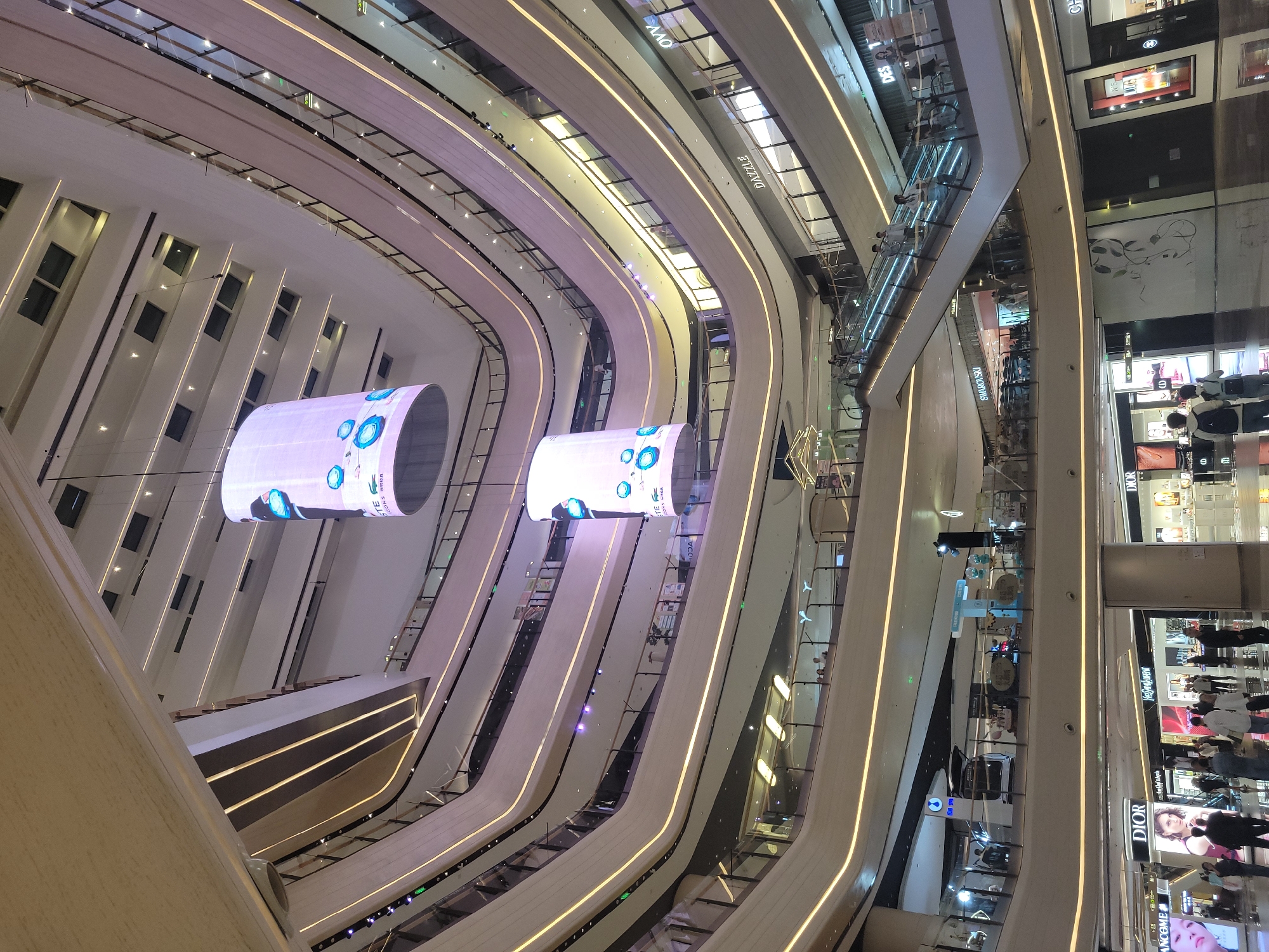 The image depicts the interior of a multi-story shopping mall in Liangjiang, China. The view is from an elevated perspective, looking down into a central atrium. The mall features curved levels, accented with linear lighting. Hanging cylindrical LED screens display graphics. Escalators are visible, connecting different levels. Retail stores with prominent brand signage such as "DIOR" line the walkways. Shoppers are present on multiple levels, browsing and walking through the mall. Windows on one side allow natural light to enter the space.