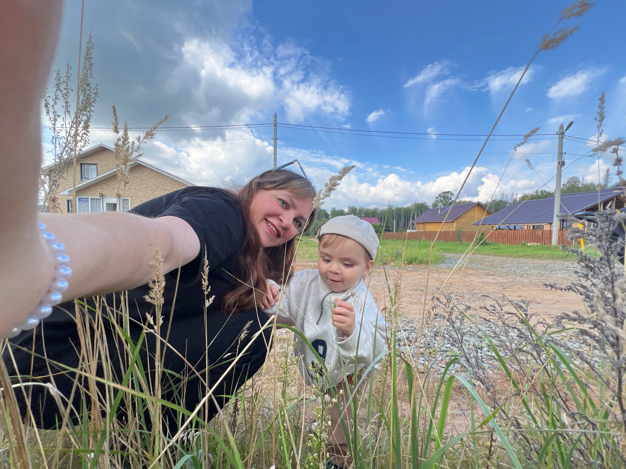 A woman and a young child are taking a selfie in tall grass. The woman is smiling at the camera, and the child is looking down at something in their hand. In the background, there are houses and trees, suggesting a rural or suburban setting. The sky is blue with scattered clouds. Based on the context, the location is likely a residential area in Noviy, Russia.