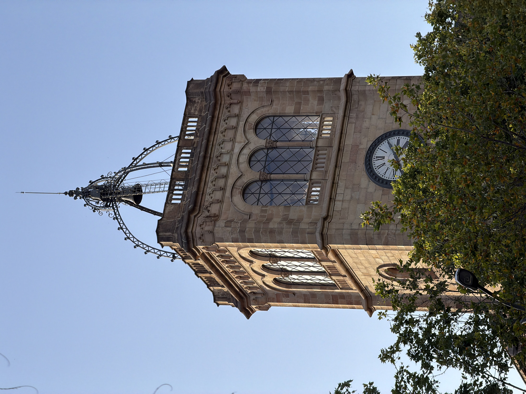 The image shows a close-up, low-angle view of a stone clock tower in Barcelona, Spain, against a clear blue sky. The tower has ornate architectural details, including arched windows with diamond-patterned panes, decorative cornices, and a balustrade near the top. Above the main body of the tower, there is an elaborate metal structure supporting bells and a spire. A large clock face with Roman numerals is visible on one side of the tower. Lush green trees with some autumn foliage partially obscure the lower portion of the tower and the foreground. A street lamp is also visible among the trees. The lighting suggests it is daytime, with sunlight illuminating the right side of the tower.