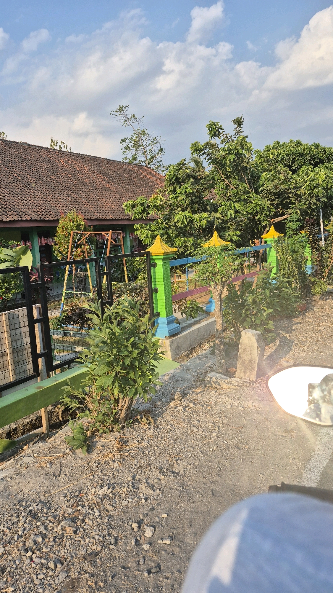 The image captures a side view of a colorful fence in front of a building with a tiled roof, possibly in a rural or suburban setting. The fence is painted with bright green posts topped with yellow caps, and has blue and pink horizontal bars. Beyond the fence, a swing set is visible, suggesting a playground area. Lush green trees and vegetation surround the property, with a clear blue sky with scattered clouds above. The foreground shows a gravelly path and a small plant. The presence of a motorcycle mirror in the lower right corner indicates the perspective is from a rider. The location is specified as Baturetno, Indonesia.