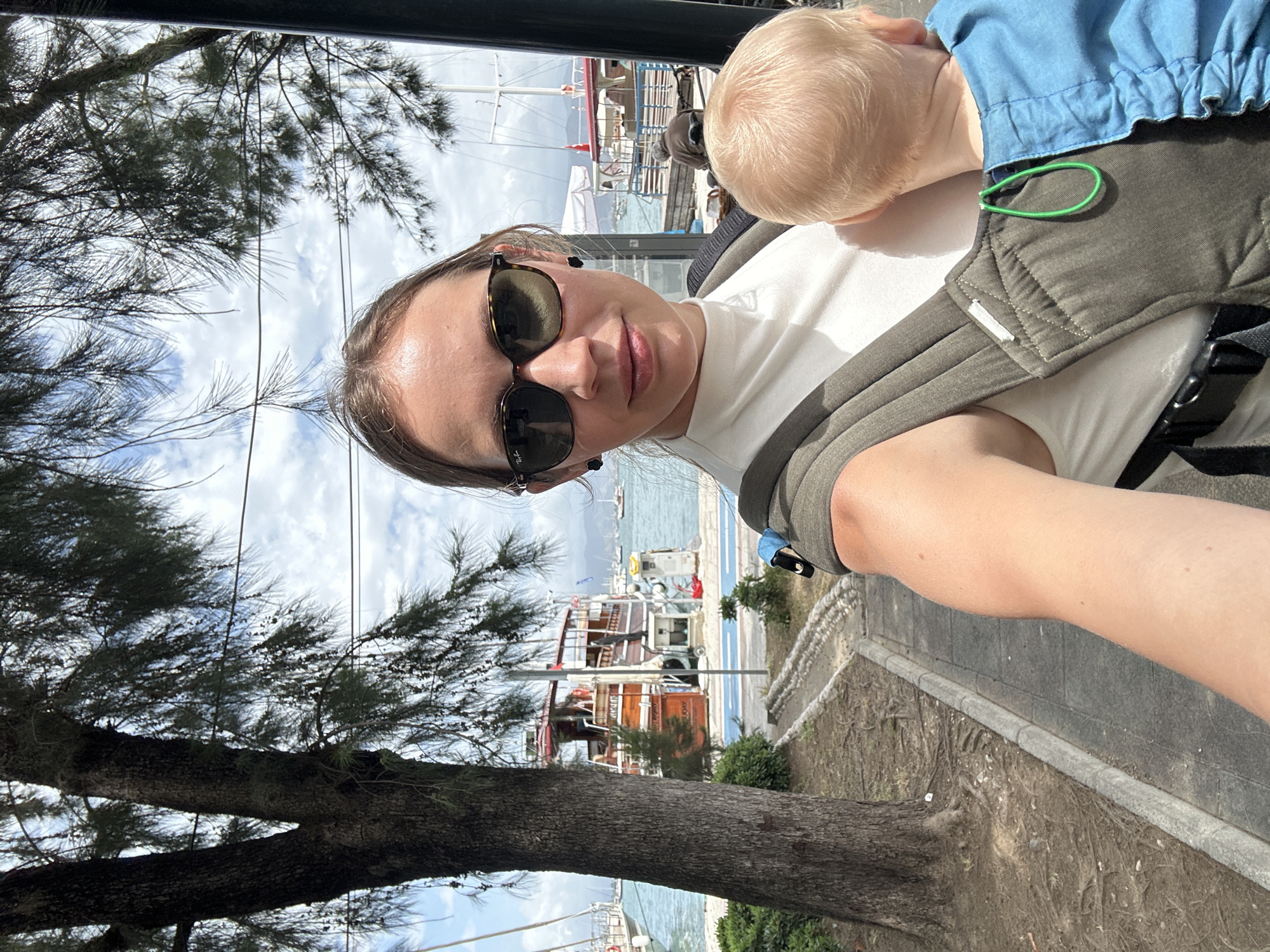 A woman wearing sunglasses is taking a selfie outdoors. She is wearing a white shirt and has a baby in a carrier on her chest. The baby is wearing a blue shirt. The location appears to be the harbor area in Fethiye, Turkey. The background features boats, a pier, and other elements typical of a coastal tourist town. A tree is partially visible on the left side of the frame.