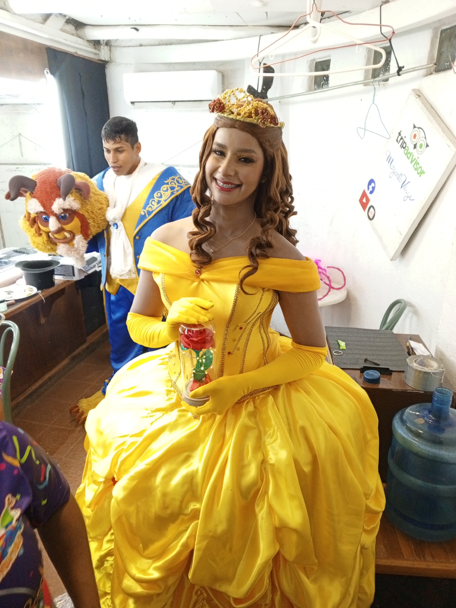 A person in a yellow Belle costume with a crown holds a light-up rose in a jar. In the background, another person holds a Beast costume head while dressed as Prince. The scene appears to be backstage, with a water jug, hangers, and a TripAdvisor whiteboard visible. The location is indicated as Margarita Village in Porlamar, Venezuela, based on the whiteboard.