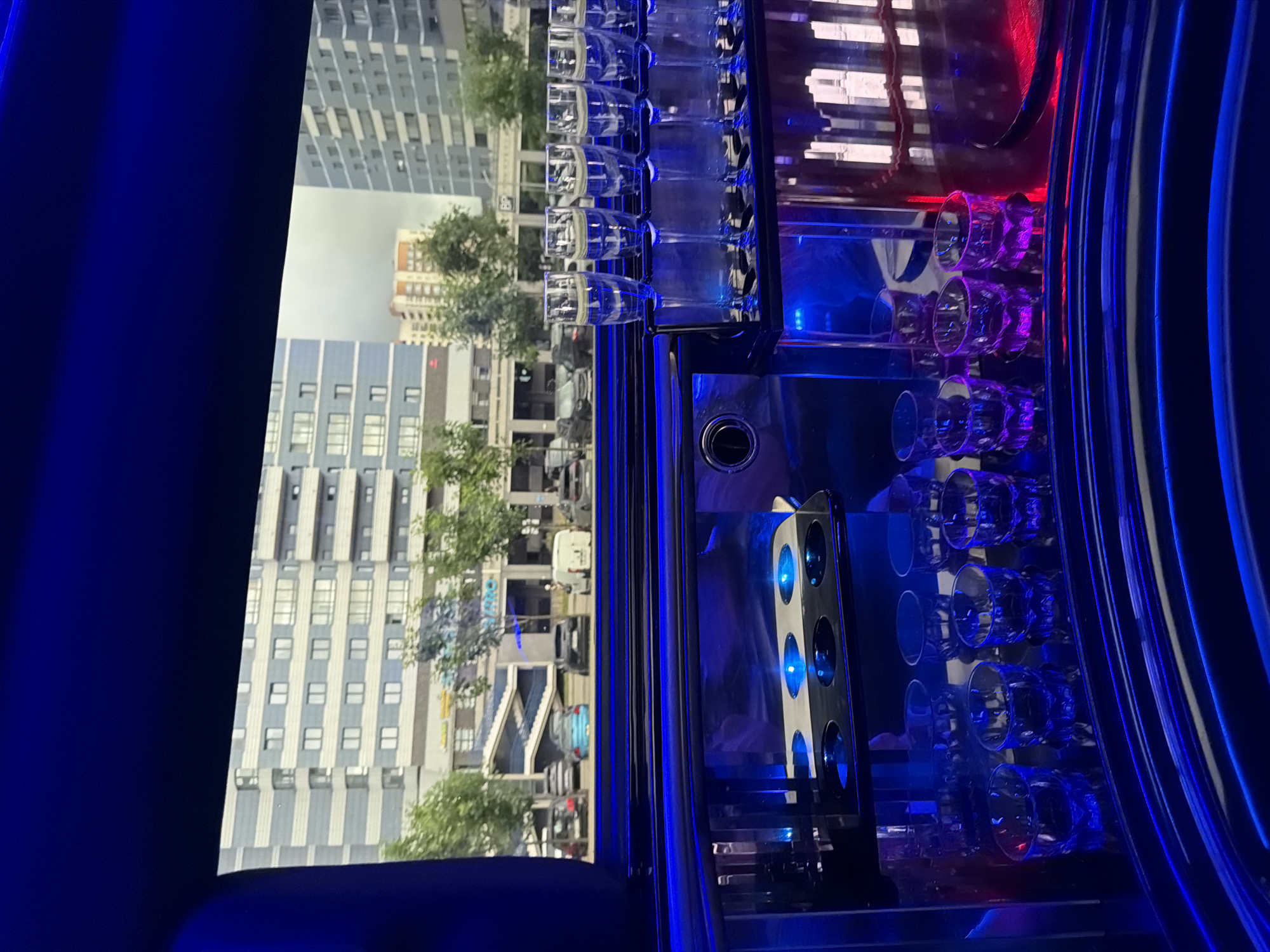 The image captures a view from inside a limousine looking out at a street scene in Minsk, Belarus. In the foreground, there is a bar area with multiple shot glasses arranged on a shelf, some of which are illuminated by blue and purple lights. Behind the bar, champagne flutes are neatly arranged in holders. The exterior view shows a row of apartment buildings with modern architecture, trees, and parked cars. One building in the background features a visible sign, potentially indicating a business or establishment. The sky appears overcast. The overall impression is one of a luxury vehicle interior juxtaposed with an urban cityscape.