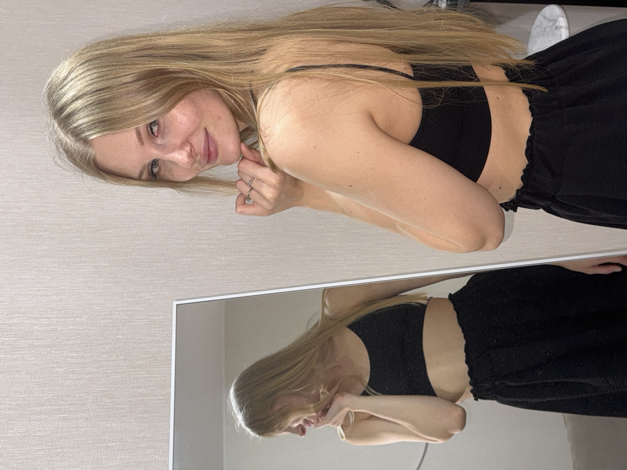 A young woman with long blonde hair is looking at the camera. She is wearing a black crop top and black shorts. She is standing in front of a mirror, and her reflection is visible. The background is a beige wall. The location is unknown, and there is no indication that it is Perm, Russia.