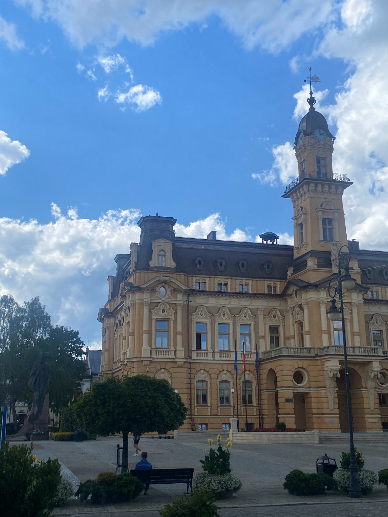 The image captures a view of the Nowy Sącz Town Hall, located in the city of Nowy Sącz, Poland. The imposing building, with its distinctive yellow facade and dark roof, features ornate architectural details, including multiple windows and decorative elements. A prominent tower with a clock face and a weather vane rises from the building. In the foreground, a paved plaza area is visible, with trees, benches, and planters. One bench is occupied by a person sitting, and another person is walking in the background. The scene is set against a backdrop of a blue sky with scattered white clouds. To the left of the building, a statue or monument is partially visible behind a tree. Flags, likely national or regional, are displayed in front of the building. A lamppost stands on the right side of the plaza. The overall impression is that of a civic or historical center in an urban setting.