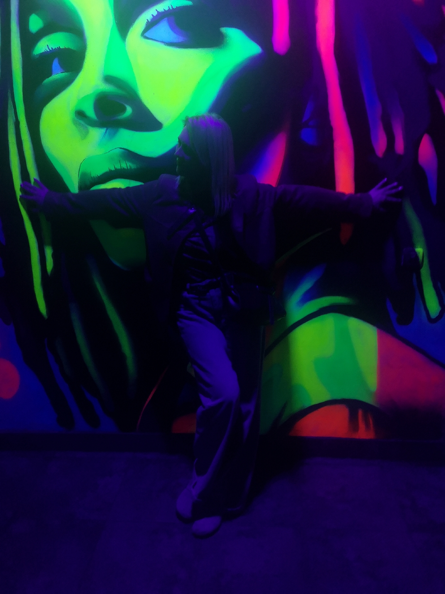 A person stands with outstretched arms in front of a vividly colored graffiti mural in Chaikovsky, Russia. The mural features a large face rendered in shades of green and blue, alongside abstract designs in pink, orange, and blue. The person is wearing dark clothing and pants, and appears to be interacting with the mural. The scene is dimly lit with predominantly blue lighting.