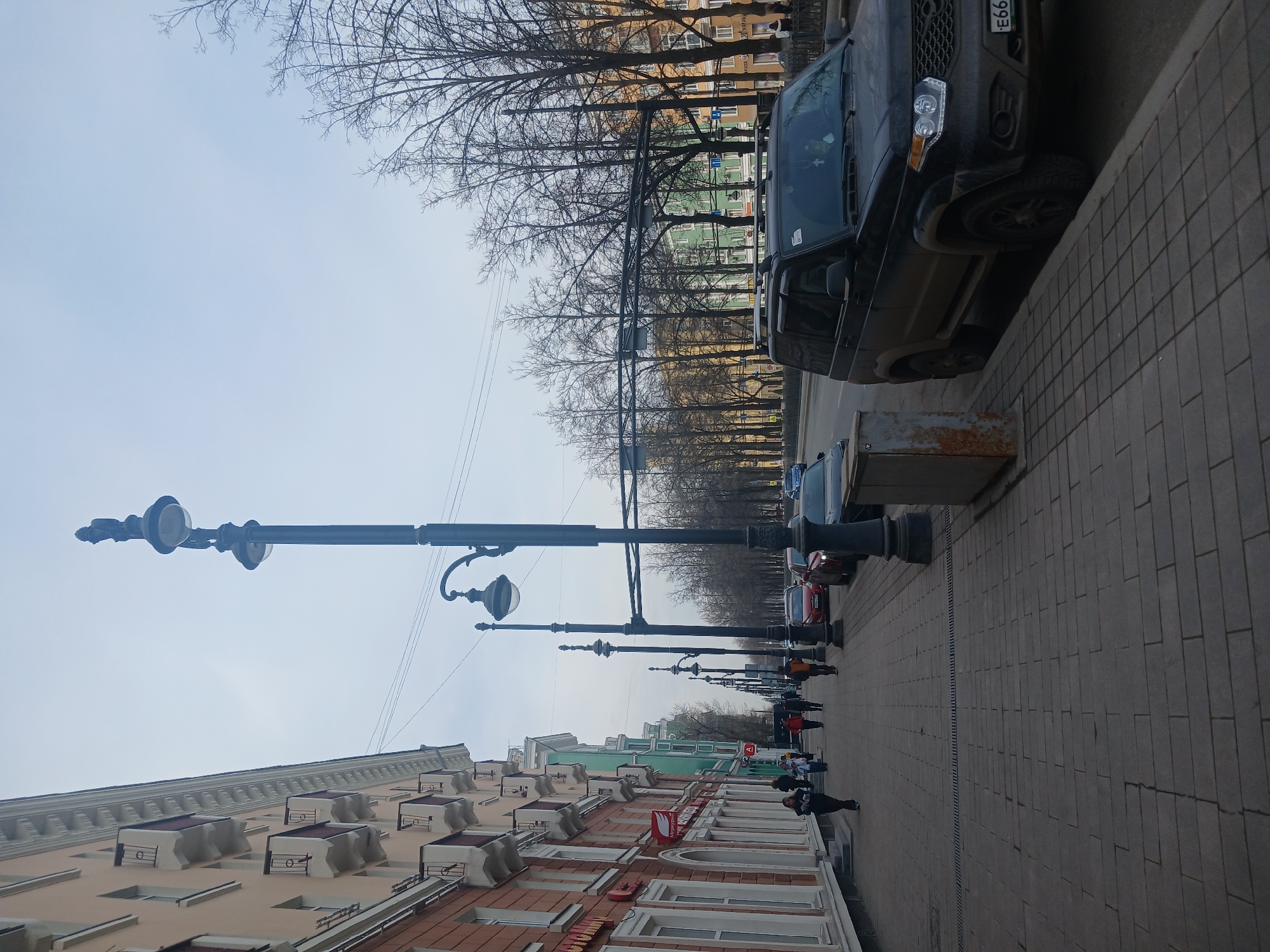 The image shows a street scene in Perm, Russia. The view is down a paved sidewalk lined with ornate street lamps and trees without leaves. Buildings are on the left, with architectural details and a mix of red, white, and beige facades. People are walking down the sidewalk, generally heading away from the viewer. Cars are parked on the right side of the street, including a dark SUV. The sky is overcast.