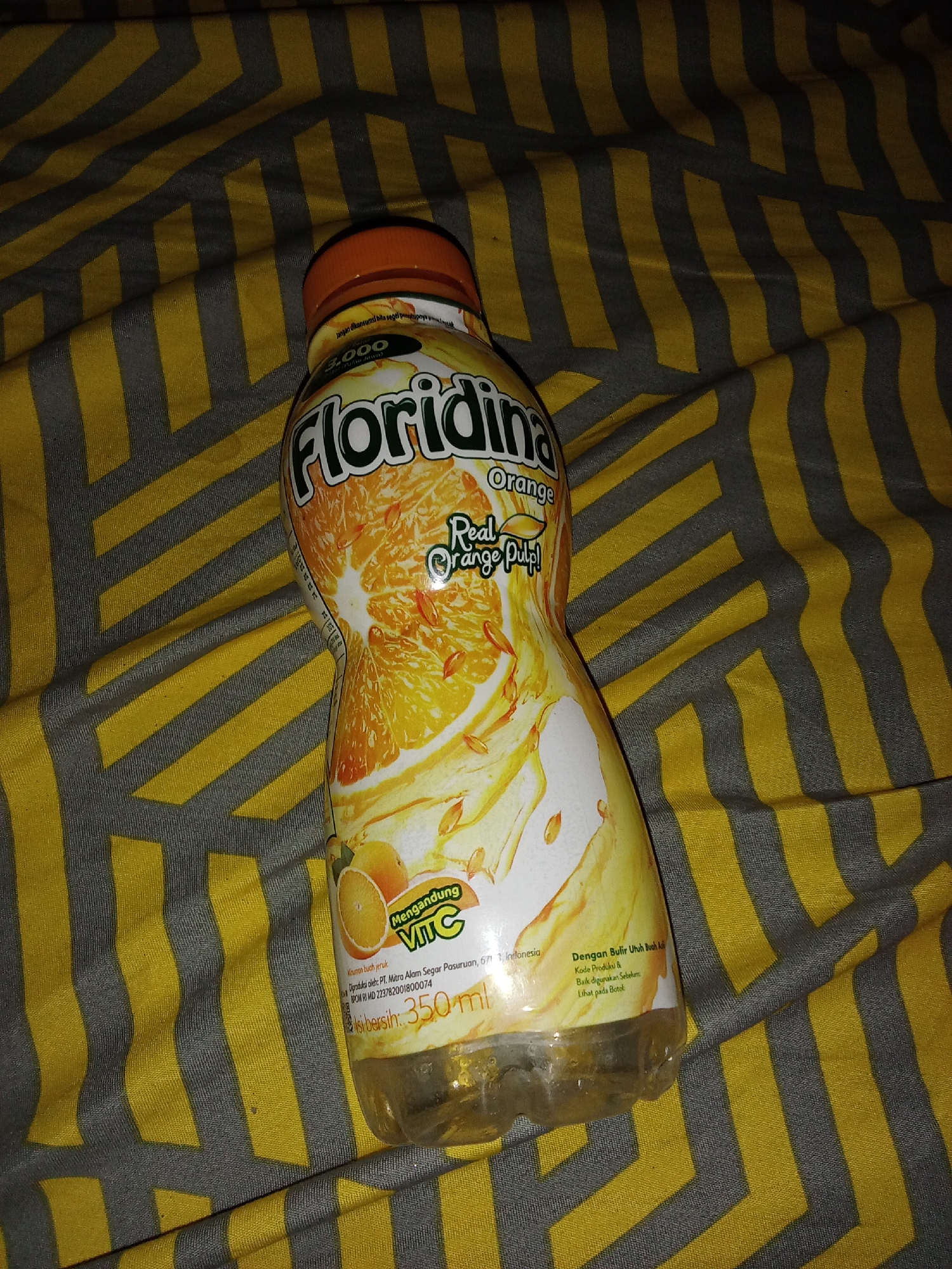 A bottle of Floridina orange juice is placed on a yellow and gray striped fabric. The bottle has an orange cap and a label featuring an illustration of oranges and the text "Floridina Orange Real Orange Pulp!". Below this, it states "Mengandung Vit C" (Contains Vitamin C) and "Kriman buah jeruk" (Orange fruit flavoring). Further text indicates it is diproduksi oleh PT. Mitra Alam Segar Pasuruan, 671 Indonesia, and lists the BPOM RI MD number. The net content is 350 ml. The fabric is likely a blanket or bedsheet. The image is taken from a top-down perspective, and the lighting suggests it was taken indoors with artificial light. There are no people visible in the image. The location is not explicitly stated to be in the city of Selowogo, Indonesia, but the product label mentions "Pasuruan, Indonesia," a city in East Java, Indonesia.