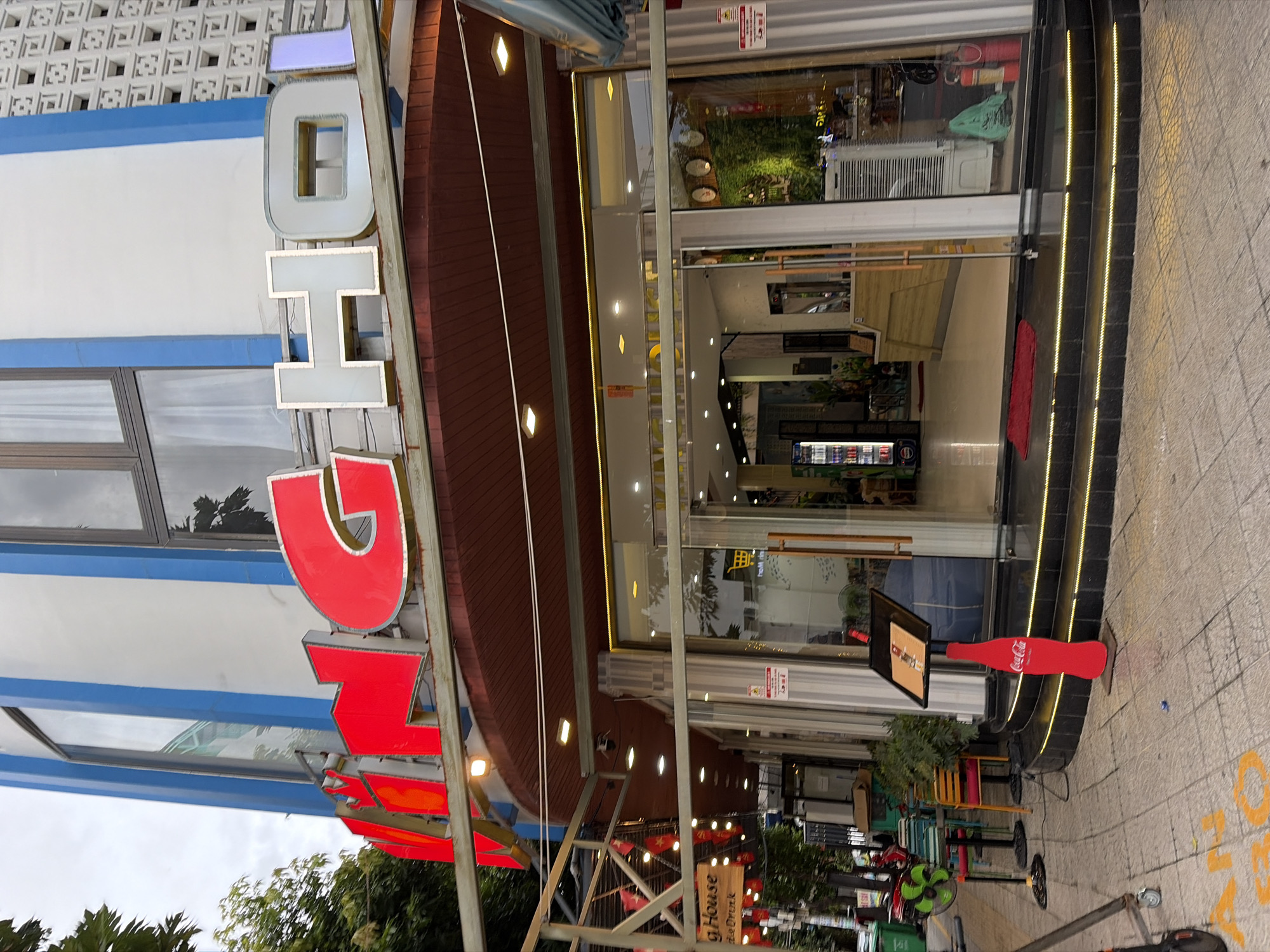 This is a medium-angle, eye-level, exterior shot of the entrance to a hotel in Đà Nẵng, Vietnam. The hotel has a modern architectural style with a white facade and blue accents. Large, red illuminated letters spelling "KING" are prominently displayed on the building, followed by white illuminated letters spelling "HOI". The entrance features a glass door and large windows that reveal a glimpse of the interior, which appears to be a reception area with plants. The building is situated on a sidewalk with yellow painted markings, and there is a red Coca-Cola bottle shaped sign on a stand near the entrance. Various objects such as chairs, tables, a fan, and a refrigerator are visible on the sidewalk, suggesting an outdoor seating area or street vendor setup. The overall scene depicts a vibrant urban environment.