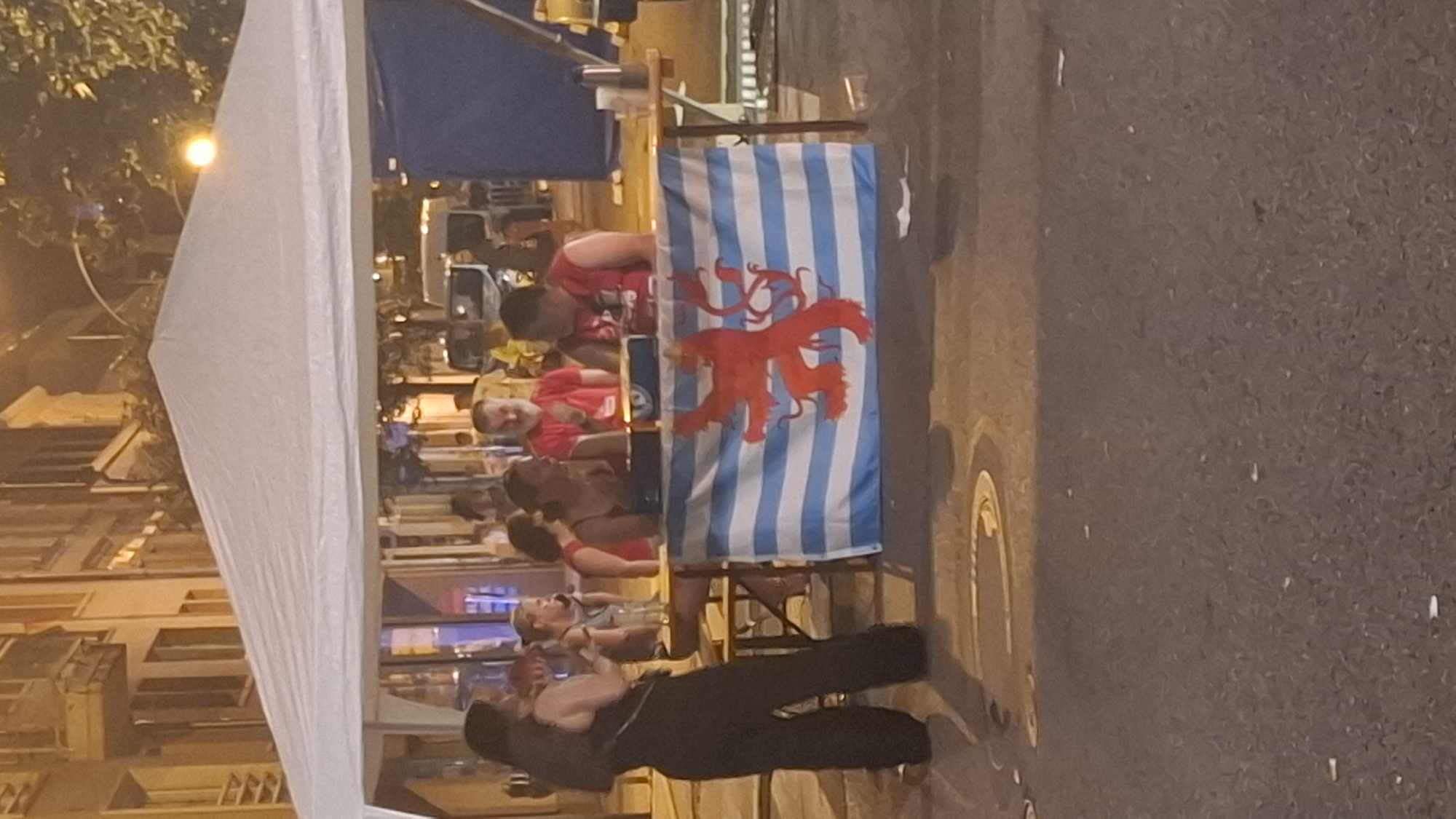 The image depicts an outdoor event likely taking place in the city of Differdange, Luxembourg, evidenced by the flag of Luxembourg displayed on a table. In the foreground, a white tent covers a table where several people are gathered. A woman in a black jumpsuit stands to the left of the table, facing the seated individuals. Three men and one woman are seated at the table, engaged in conversation or activity. The man on the far right, wearing a red shirt, appears to be interacting with the woman next to him. In the background, buildings and streetlights suggest an urban setting, possibly a street or plaza, with trees and parked vehicles visible. The lighting indicates the event is taking place during the evening or night. The flag of Luxembourg, featuring horizontal stripes of white and light blue with a red lion rampant in the center, is prominently displayed on the front of the table.