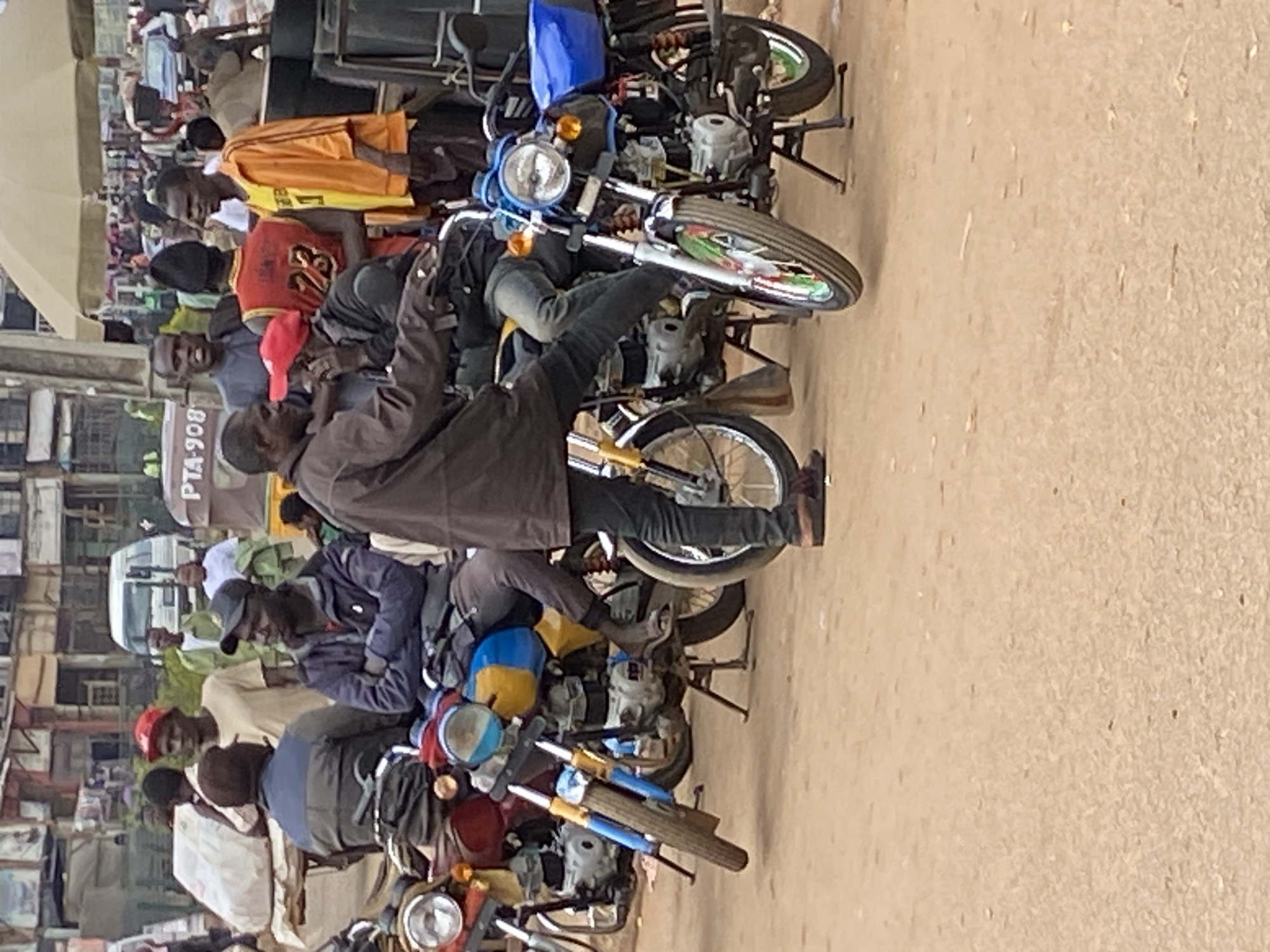 The image captures a bustling street scene in Bukuru, Nigeria, likely at a marketplace. Several men are present, some standing and others seated on motorcycles. In the foreground, a group of men are gathered around motorcycles. One man, wearing a dark jacket and jeans, is seated on a motorcycle with a blue and yellow cover, while another man in a dark jacket is standing nearby, facing him. A third man, in an orange jacket, stands to the right, looking away from the camera.
Further back, several other individuals are visible, some of whom appear to be vendors or shoppers. The environment suggests a busy urban setting with buildings and possibly stalls in the background. The ground is unpaved, typical of many streets in Nigerian cities. The lighting indicates it is daytime, likely mid-morning or afternoon. The overall impression is one of everyday life in a busy Nigerian marketplace.