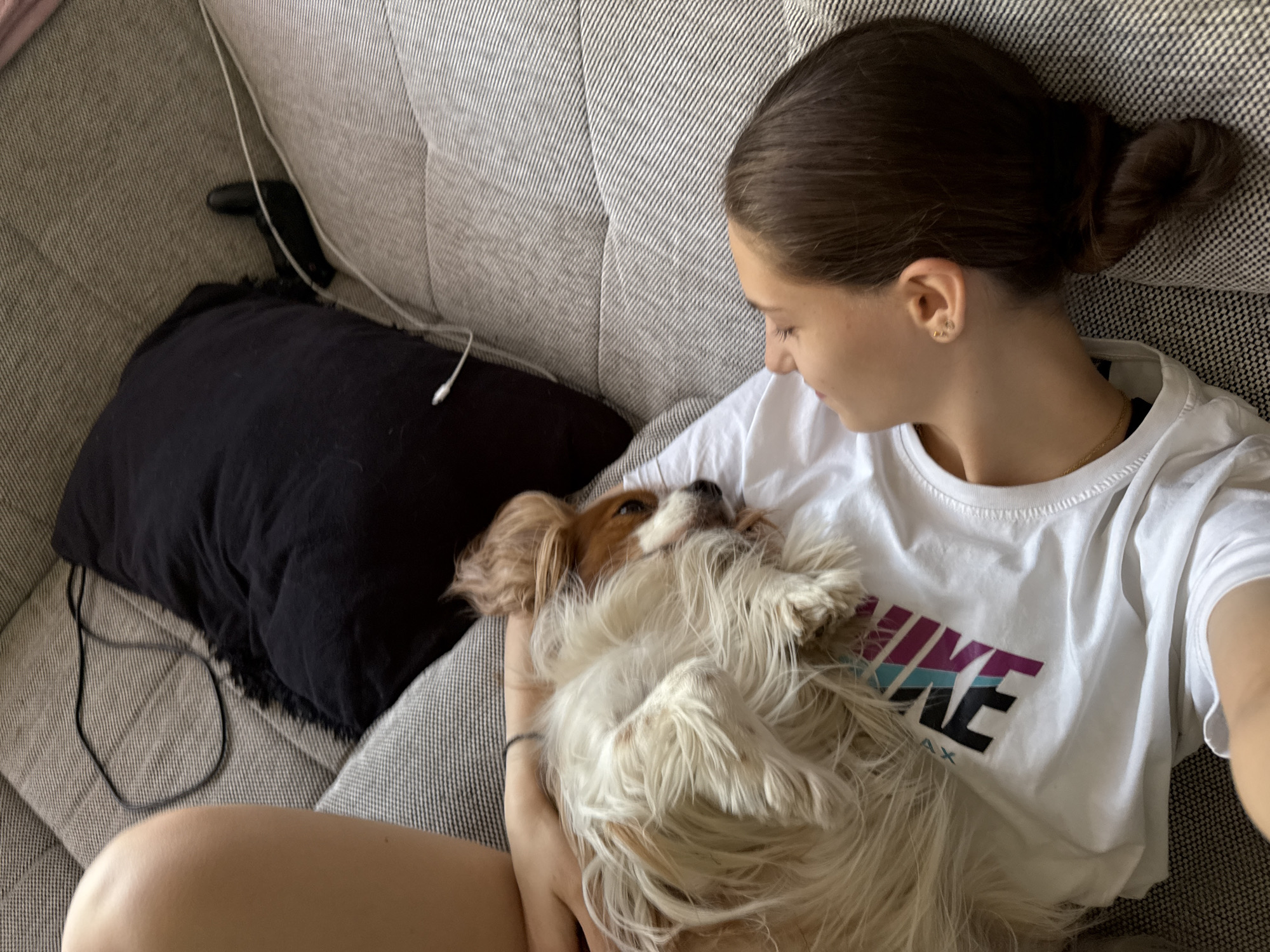 A young woman with brown hair in a bun is sitting on a gray textured couch, holding a Cavalier King Charles Spaniel dog. She is wearing a white Nike t-shirt. A black pillow is visible to the left. A black object and a white charging cable are on the couch near the pillow. The location appears to be an interior setting. This image was not taken in the city of Sofia, Bulgaria.