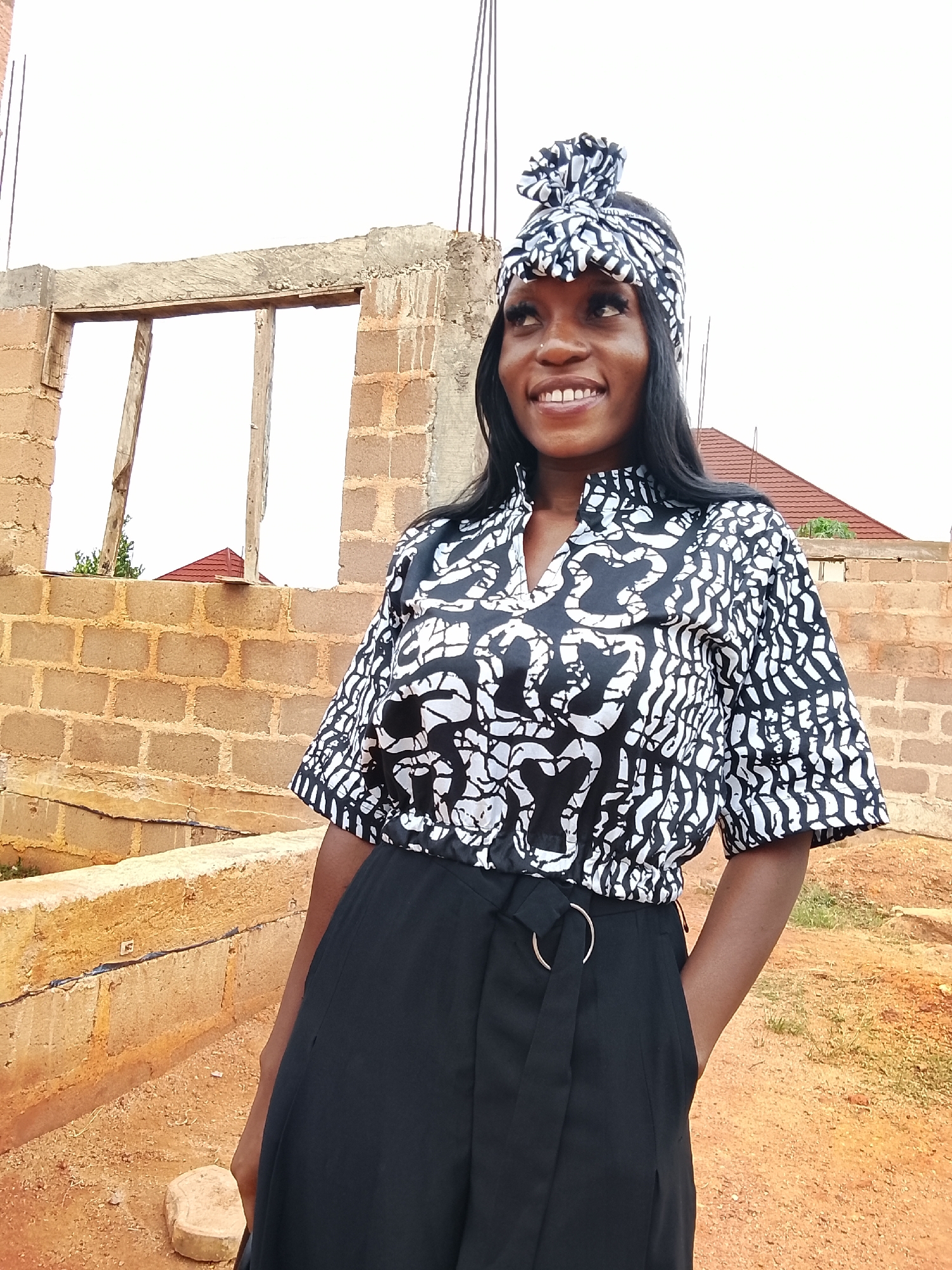 A young woman stands in front of a partially constructed brick building in Barikin Sabon Gidan Kanat, Nigeria. She wears a black and white patterned top and matching headwrap, paired with black pants. Her right hand is in her pocket, and she is smiling and looking to her left. The building behind her has open window frames with wooden supports. In the background, there is a glimpse of a red-roofed structure and an overcast sky. The ground is reddish-brown dirt.