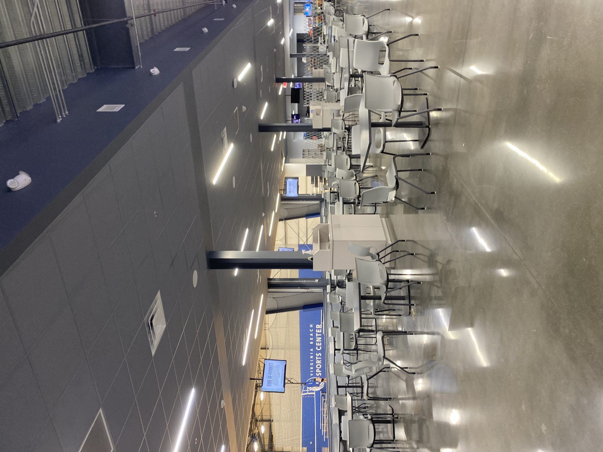 The image depicts an interior view of the Virginia Beach Sports Center, located in Virginia Beach, United States of America. The scene is dominated by rows of empty tables and chairs, set on a polished concrete floor that reflects the overhead lighting. The tables are square and dark-colored, with matching dark-colored chairs featuring thin metal legs.
In the background, a large blue wall with white lettering reads "VIRGINIA BEACH SPORTS CENTER". Above this wall, a screen displays "CODE OF CONDUCT". Further in the background, a sports netting structure is visible, suggesting the presence of courts or fields. The ceiling is a dark gray drop ceiling with numerous fluorescent light fixtures. There are also several dark-colored support columns spaced throughout the area. The overall environment appears clean and well-maintained, designed for a public facility. There are no people visible in the image.