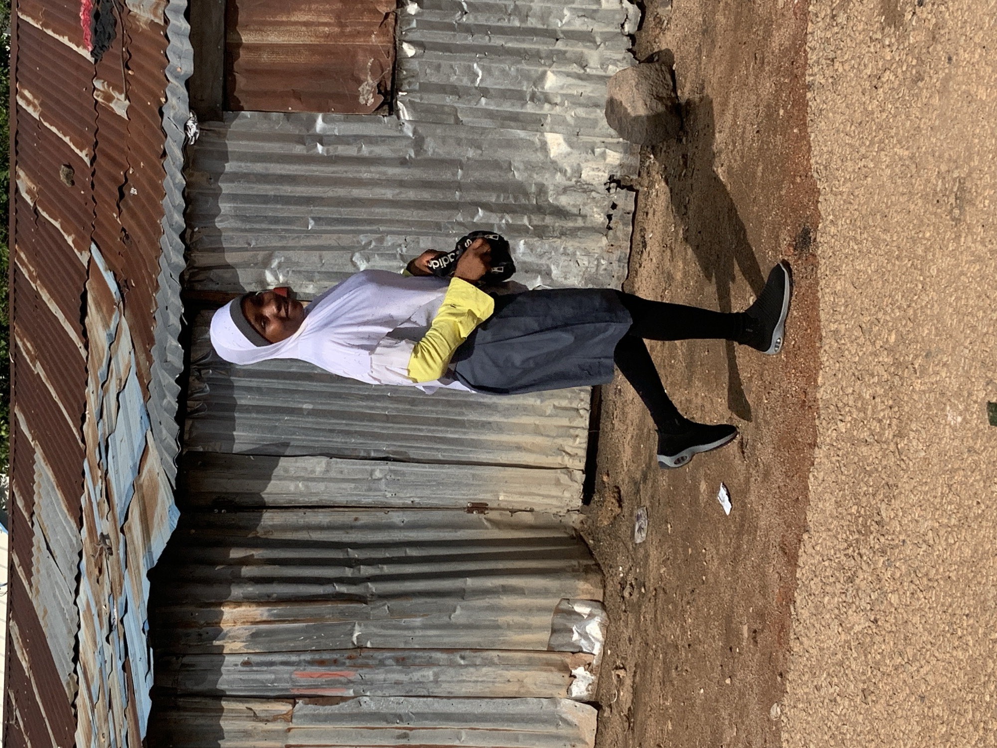 A young woman wearing a white hijab, a white top with bright yellow sleeves, and a dark blue skirt walks along a dirt path in front of a corrugated metal structure. She is carrying a black bag with white lettering. The structure appears to be a makeshift building, with rust and some damage visible on its metal exterior and roof. The ground is composed of dirt and gravel. The scene is lit by natural sunlight, casting shadows. The image appears to have been taken outdoors, possibly in a residential or informal settlement area within the city of Jos, Nigeria, given the typical architectural style of such areas.