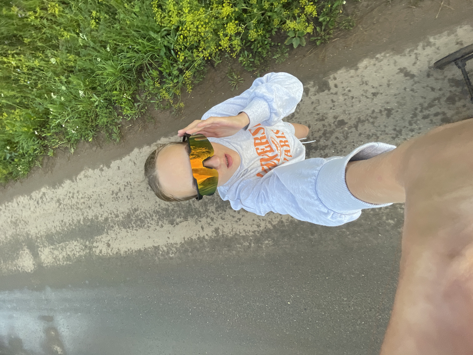 A high-angle, first-person perspective selfie is taken from the perspective of someone on a bicycle, looking down and slightly forward. The scene is outdoors, with a wet asphalt or dirt path occupying the lower half of the frame. To the right of the path, tall green grass and yellow wildflowers are visible, suggesting a rural or park-like setting. In the upper portion of the frame, a young woman is visible, wearing a light blue hoodie with orange lettering that appears to say "UNIVERSITY PARIS". She has large, reflective sunglasses on and is holding one hand up to the side of her sunglasses, as if adjusting them or shielding her eyes. Her expression is one of mild surprise or exertion. The image is captured on a cloudy or overcast day, as indicated by the diffused light. The location is identified as Podshivalovo, Russia.