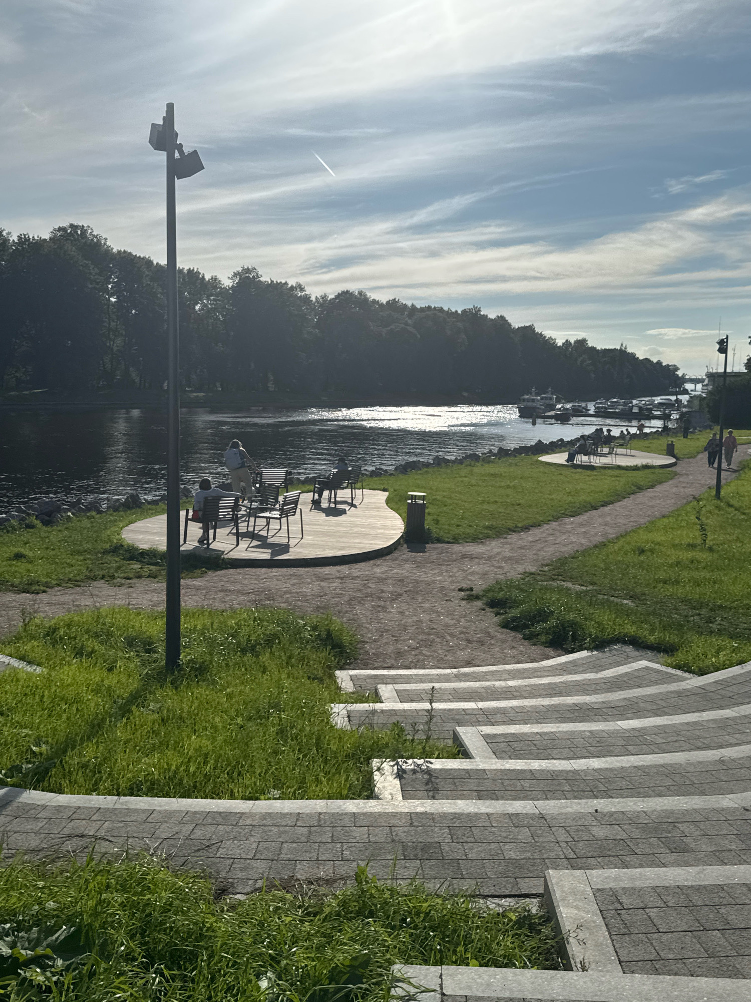 The image depicts a scenic view of the waterfront in Saint Petersburg, Russia, likely in an area with recreational amenities. In the foreground, a set of tiered stone steps curves towards a paved pathway that winds through lush green grass. A tall lamp post with two lights stands prominently to the left of the steps, casting a shadow on the grass.
Further back, a circular wooden deck features a table with several chairs, where a few individuals are seated. Beyond this area, a calm body of water, possibly a river or bay, reflects the bright sunlight. Along the opposite bank of the water, a dense line of trees creates a dark, verdant backdrop. In the distance, several boats are moored at a dock, suggesting a marina or harbor. The sky above is a soft blue, streaked with wispy white clouds, and a faint contrail from an aircraft is visible. Two people are walking along the pathway on the right side of the frame, approaching the boats. The overall impression is one of a peaceful, well-maintained public space during daylight hours.