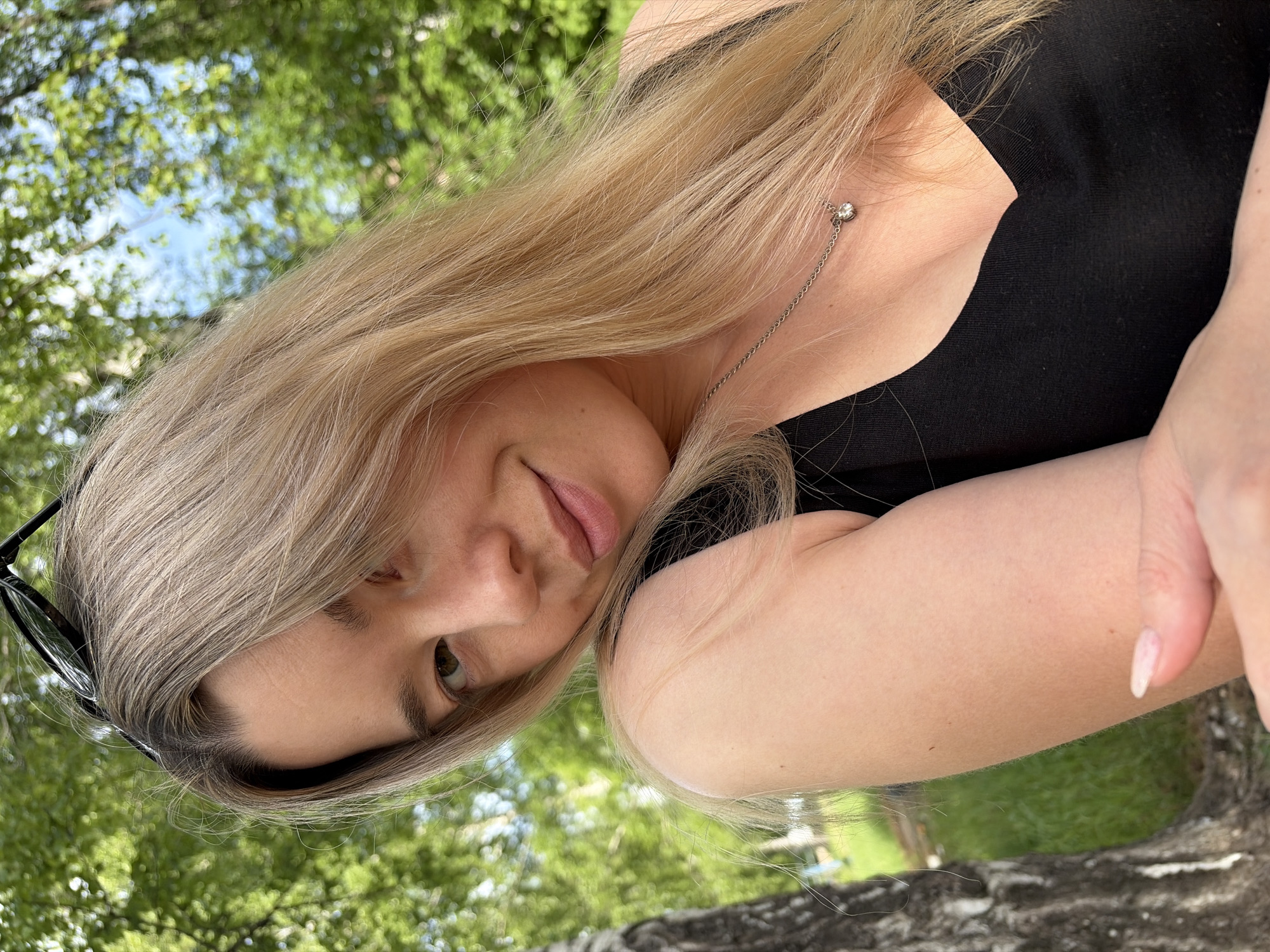 A woman with long, blonde hair is pictured outside. She is wearing a black top and a necklace with a small pendant. A pair of sunglasses is positioned on her head. The background consists of green trees and foliage, suggesting a park or green space. It is not possible to determine her location in Chaikovsky, Russia from the image provided.