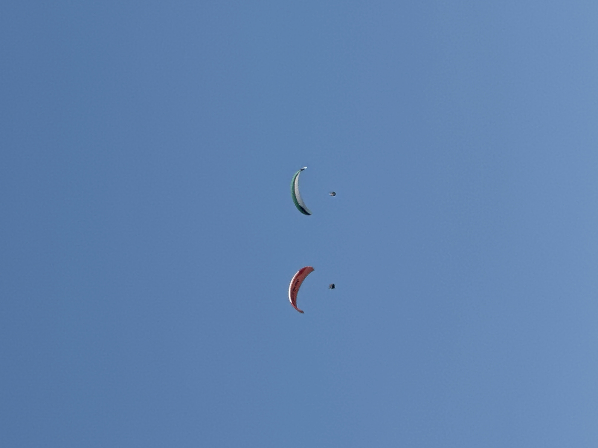 The image shows two paragliders in a clear blue sky, with no visible land features or city elements suggesting Boreti, Montenegro. One paraglider, colored red and white, is to the left, and another paraglider, green and white, is to the right. Both paragliders appear to be in mid-flight.
