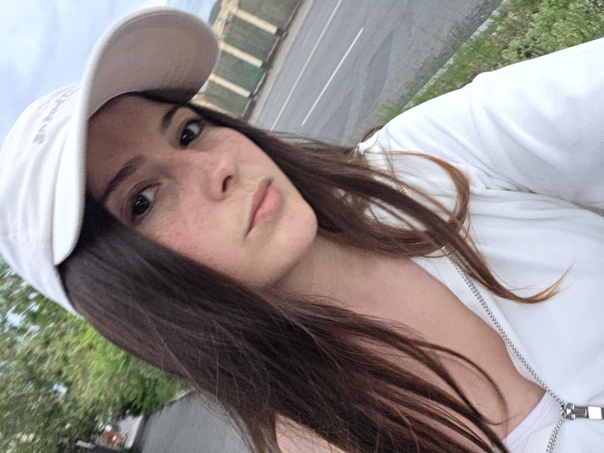 This is a selfie of a young woman wearing a white baseball cap and a white zip-up hoodie. She has long, dark brown hair that falls around her shoulders and chest. Her eyes are open and she is looking towards the camera with a neutral expression. She has freckles across her nose and cheeks. The background is blurred, but appears to be an outdoor setting with a road and some greenery visible. The lighting suggests it is daytime, possibly overcast due to the soft light. There is some text visible on the baseball cap, but it is too blurry to read. The woman's clothing is simple and casual.