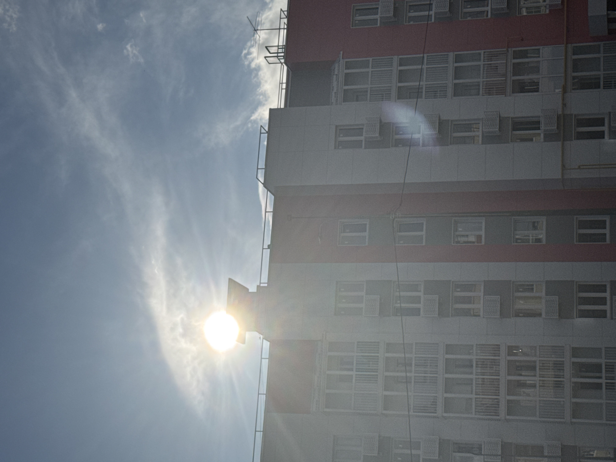 The image captures a modern apartment building in Volokolamsk, Russia, under a bright, partly cloudy sky. The sun is high and bright, creating lens flares that streak across the frame and partially obscure the upper portion of the building. The building itself is constructed with sections of light gray and a vibrant pinkish-red. Numerous windows and balconies are visible, many with white railings. Air conditioning units are mounted below some of the windows. A telecommunications antenna is mounted on the roof of the building on the right side. The scene appears to be during daylight hours, likely morning or afternoon, given the sun's position and intensity. There are no people or vehicles immediately visible in the frame. The overall impression is of a residential area bathed in strong sunlight.
