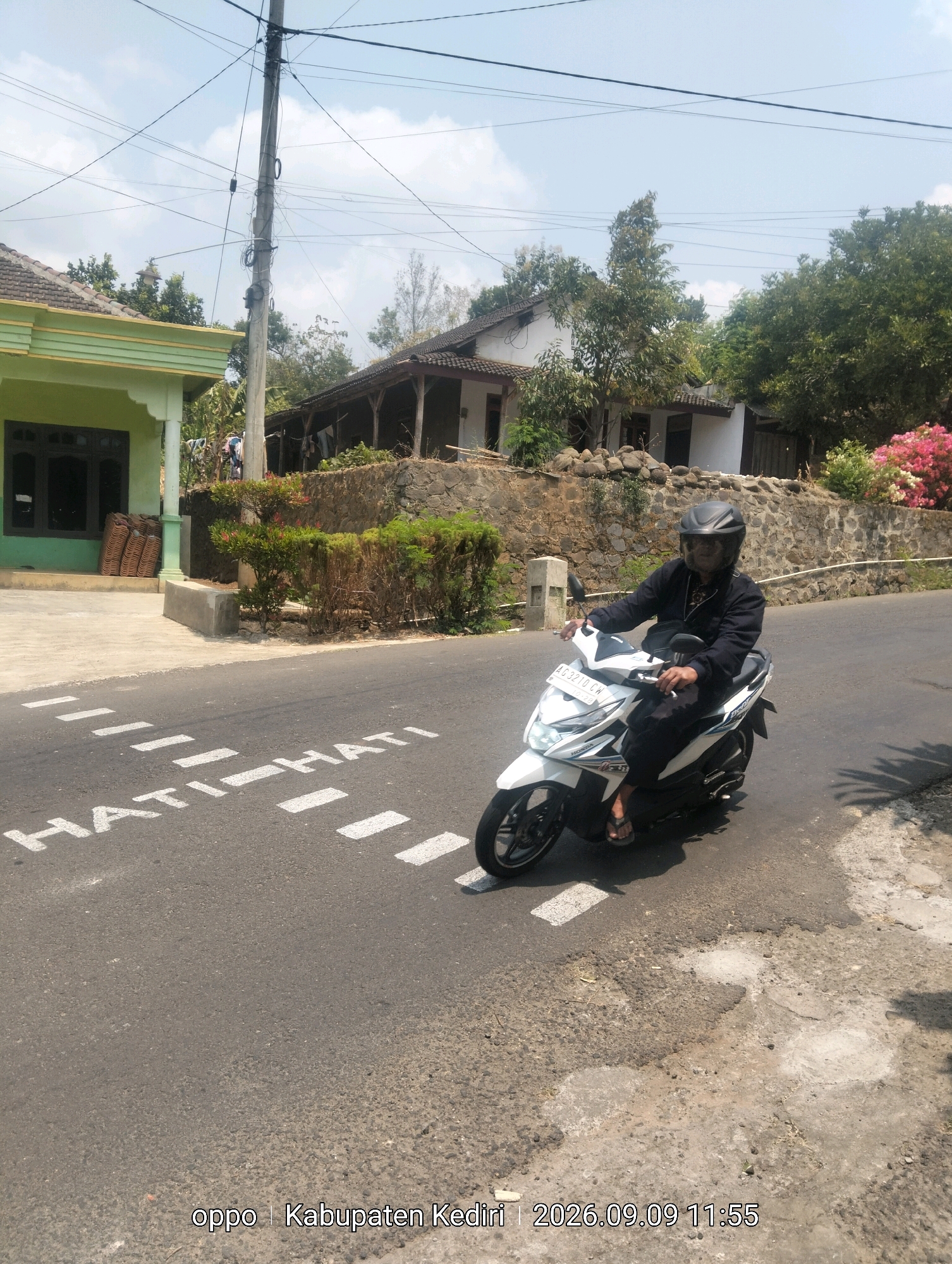 An outdoor, eye-level, medium shot captures a person riding a white Honda scooter on an asphalt road. The rider, wearing a black jacket and helmet, is positioned in the center of the frame. White dashed lines and the white text "HATI HATI" (meaning "Be Careful" in Indonesian) are painted on the road in the foreground, indicating a pedestrian crossing. To the left of the road is a light green house with a dark-trimmed window. Behind the house and across the road, a stone wall borders a residential area with a white house and some trees. The sky is clear blue with scattered clouds. The timestamp on the image reads "2026.09.09 11:55" and the location is identified as "Kabupaten Kediri."