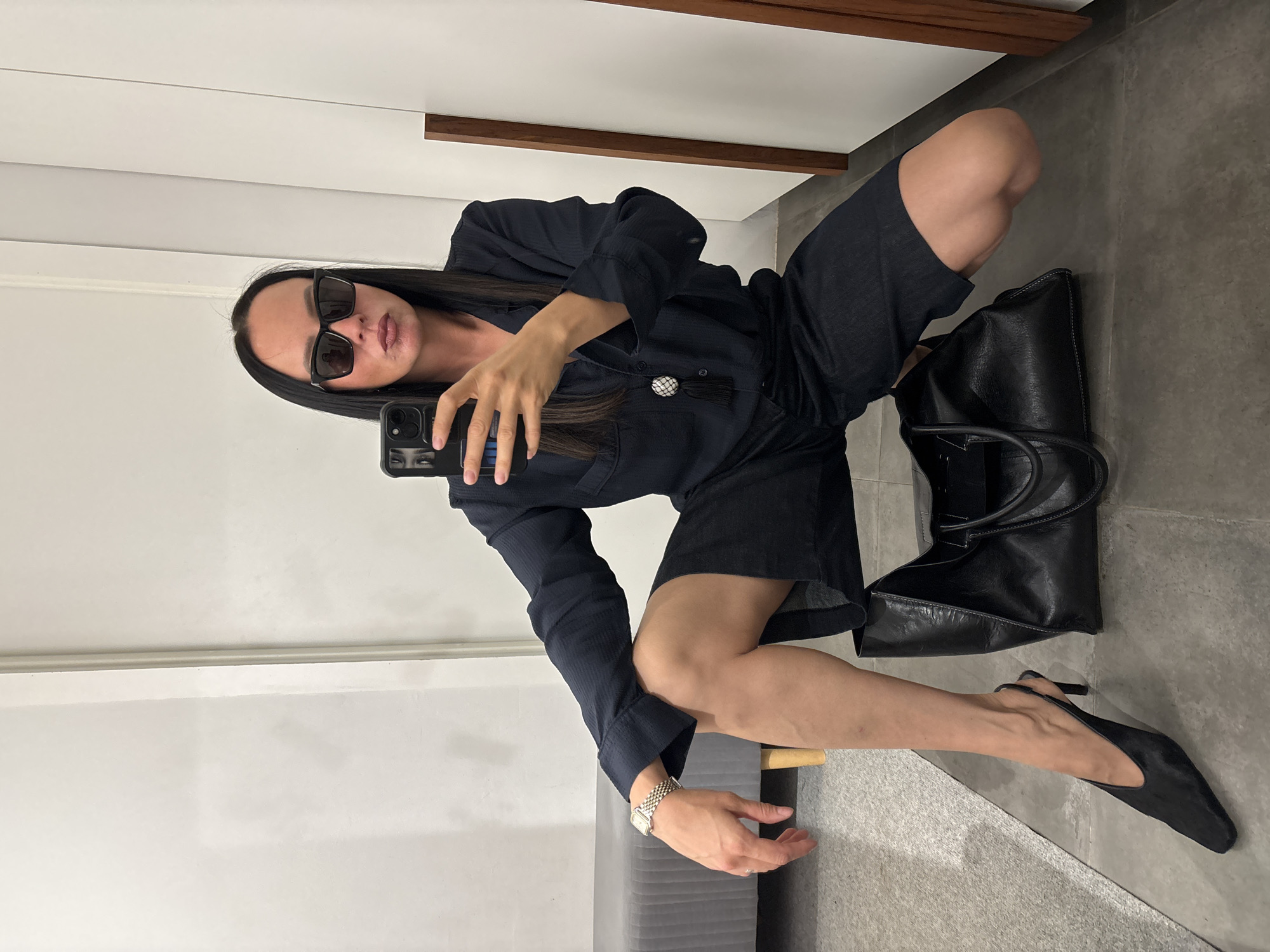 A person is squatting while holding a phone, taking a mirror selfie. The person is wearing a dark blue, short-sleeved jumpsuit, dark sunglasses, and a silver watch. A black leather bag is placed on the ground next to the person, and they are wearing black heels. The scene appears to be indoors, with white walls and a gray floor. A bench or seat is partially visible in the bottom left corner. No specific details suggest the location is Chaikovsky, Russia.