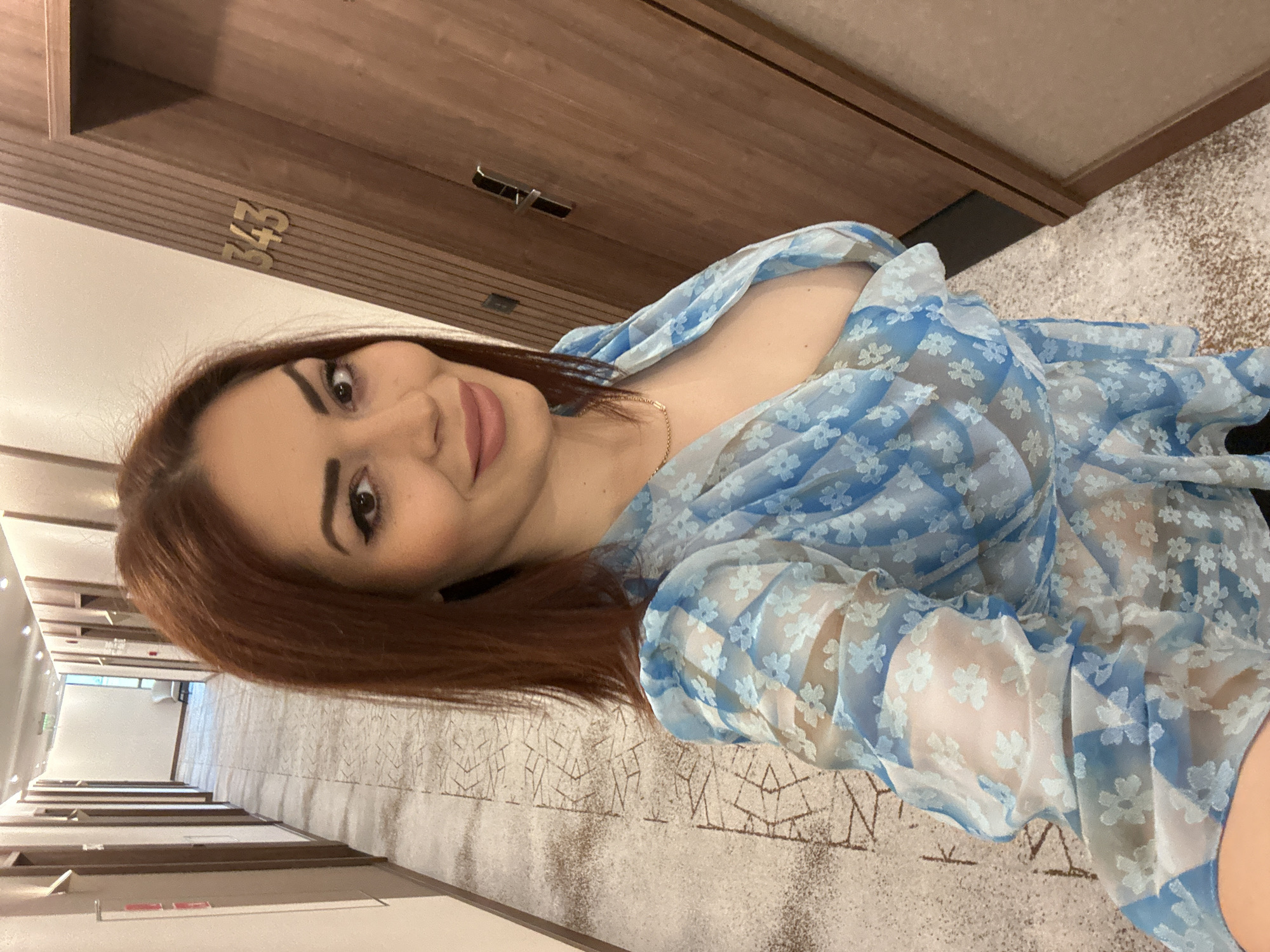 A woman with red hair is standing in a hotel hallway. She is wearing a sheer blue floral print top. The hallway has a beige patterned carpet and wooden doors with room numbers. The room number "343" is visible on the door behind her. The hallway stretches into the distance, with multiple doors and a glimpse of a window at the end. The location cannot be determined based on the image alone, nor whether it is in Kazanlak, Bulgaria.