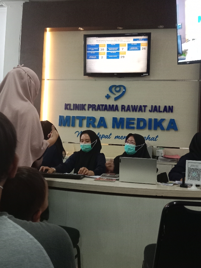 The image captures the interior reception area of Klinik Pratama Rawat Jalan Mitra Medika in Gladaksari, Indonesia. Three female staff members, all wearing head coverings and light green surgical masks, are positioned behind a light-colored counter. The central staff member has an open silver laptop in front of her. On the counter's right side, a white stand displaying a QR code is visible. Behind the counter, the clinic's name and a heart-plus logo are prominently displayed in blue lettering on a light wall, framed by vertical LED light strips. Above this, a wall-mounted television displays a patient queue system with various numbers including 30, 7, 26, 2, and 29. A second, partially visible television is mounted to the right. In the foreground, a person wearing a light pink head covering stands facing the counter, while two additional individuals are seated with their backs to the camera, indicating a waiting area.