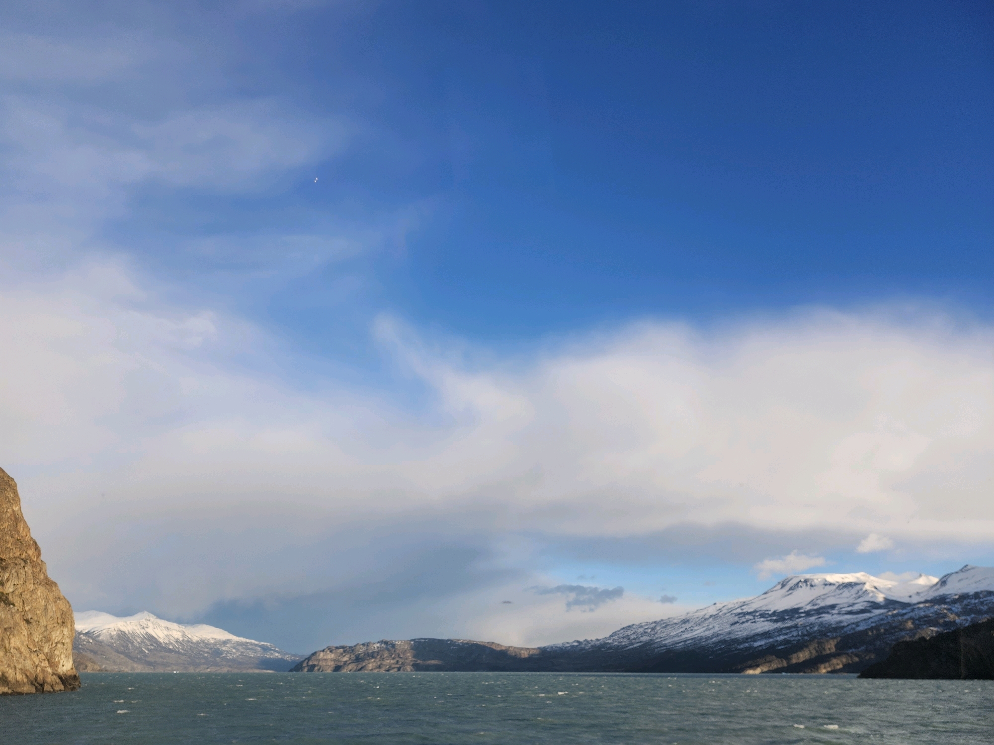 The image shows a wide shot of a body of water with snow-capped mountains and rocky cliffs in the background. The sky is a bright blue with scattered white clouds. The water is a deep green and appears choppy with small waves. On the left side of the image, a large, rocky cliff face occupies the foreground. In the distance, snow-covered mountains stretch across the horizon, with some rocky outcrops visible among them. The scene suggests a remote, natural landscape.