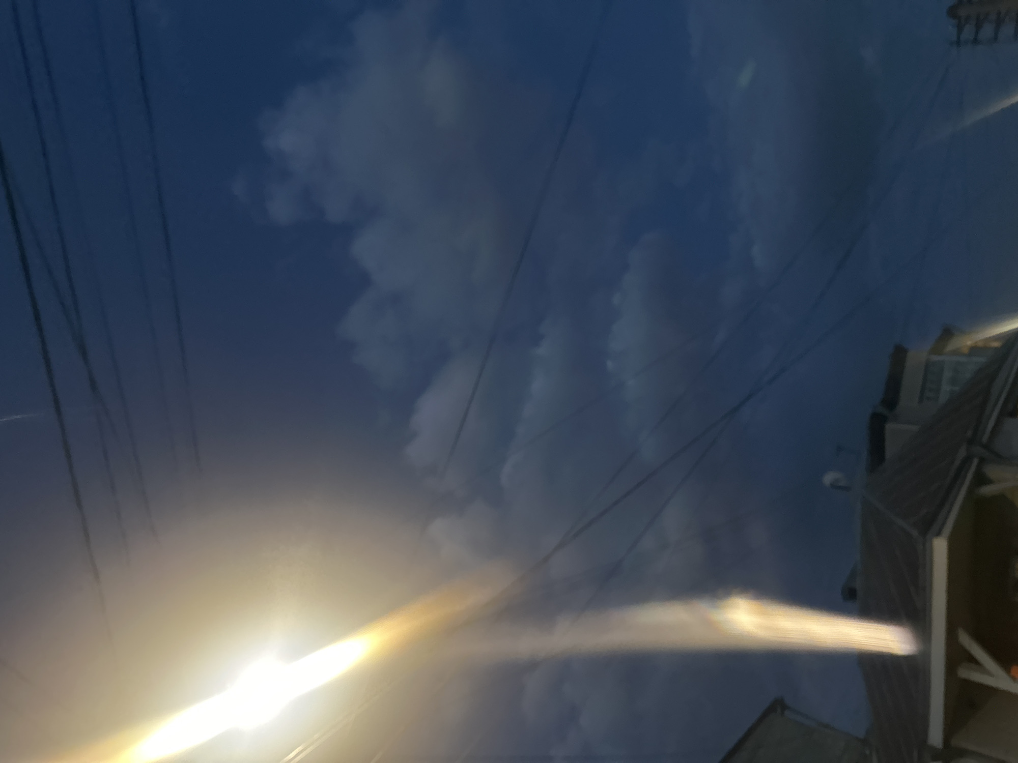 The image, taken in Yerevan, Armenia, shows a night scene with an upward view. The sky is a deep blue with patches of white clouds. Several electrical wires crisscross the sky. In the lower left corner, there's a bright, diffused light source, possibly a streetlight, creating a lens flare effect across the image. To the right, the corner of a building is visible, with its roof and upper stories partially illuminated.