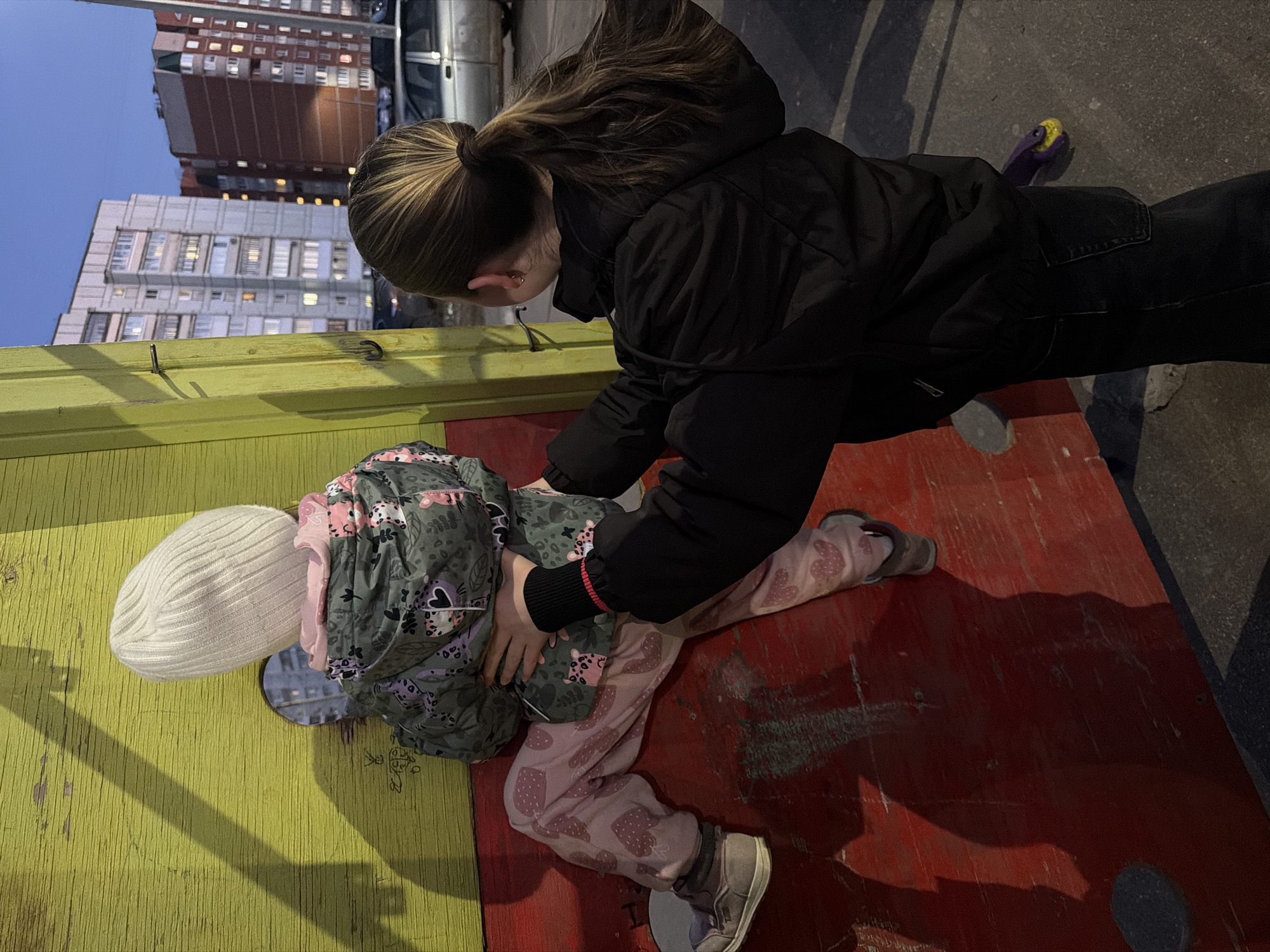 A child is in a wooden pillory, with their head through a hole on the left, and their body on the right. The pillory is painted with a yellow board on the left and red board on the right. An older child in a black hooded jacket is adjusting the child in the pillory. The child in the pillory is wearing a white beanie, a green patterned jacket, and pink patterned pants with gray shoes. In the background are apartment buildings. The location is Chaikovsky, Russia.