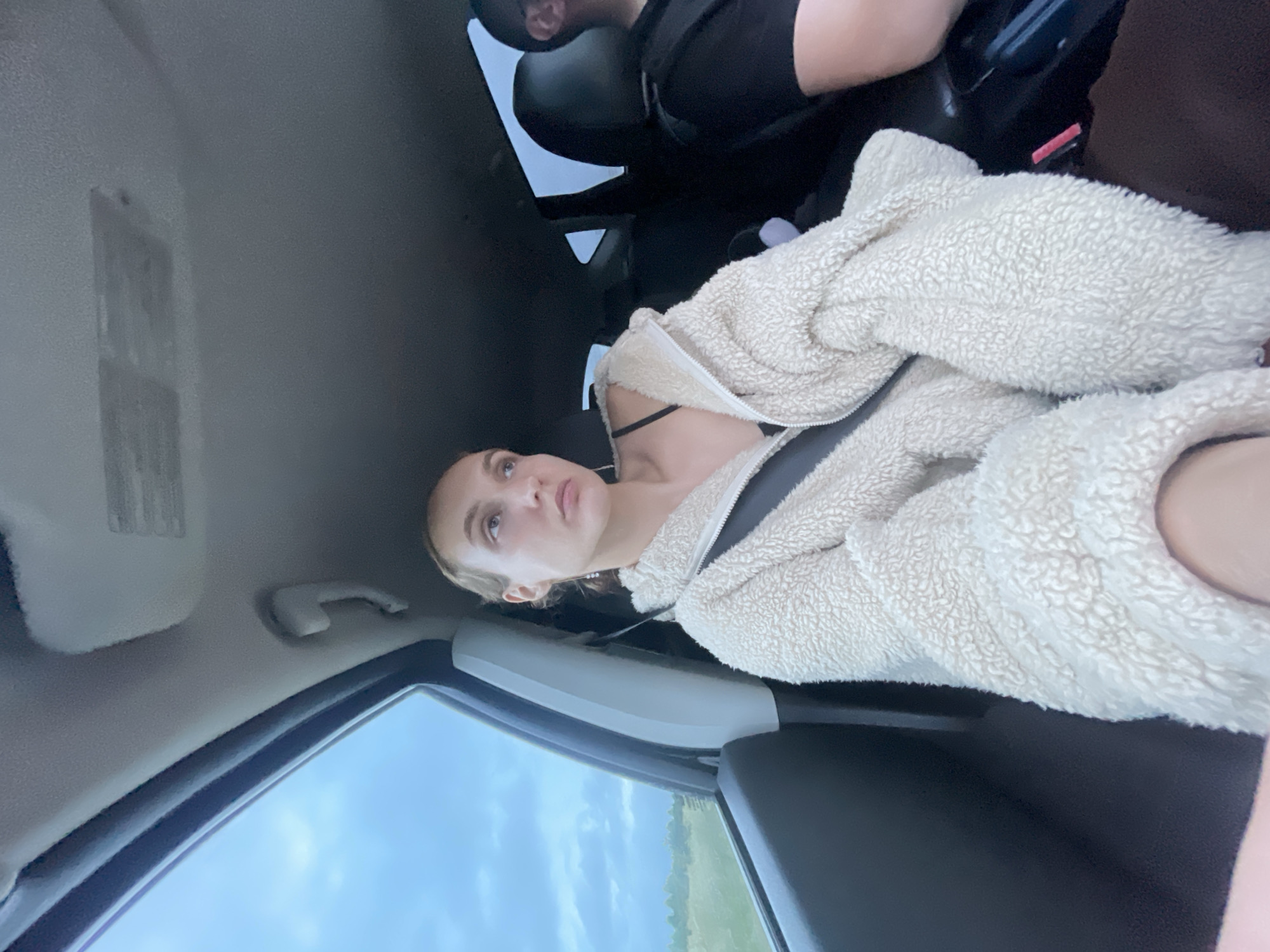 A medium shot captures a young woman with light brown hair pulled back, wearing a cream-colored fuzzy jacket and a black bra, seated in the passenger seat of a vehicle. Her gaze is directed to her right, with a thoughtful expression. A seatbelt is fastened across her chest. The interior of the car is visible, including the ceiling and part of the driver's seat and shoulder in the background. Through the car window, a cloudy, overcast sky and a flat, grassy landscape can be seen, suggesting a rural or roadside setting. The location is identified as Meshcheryaki, Russia.