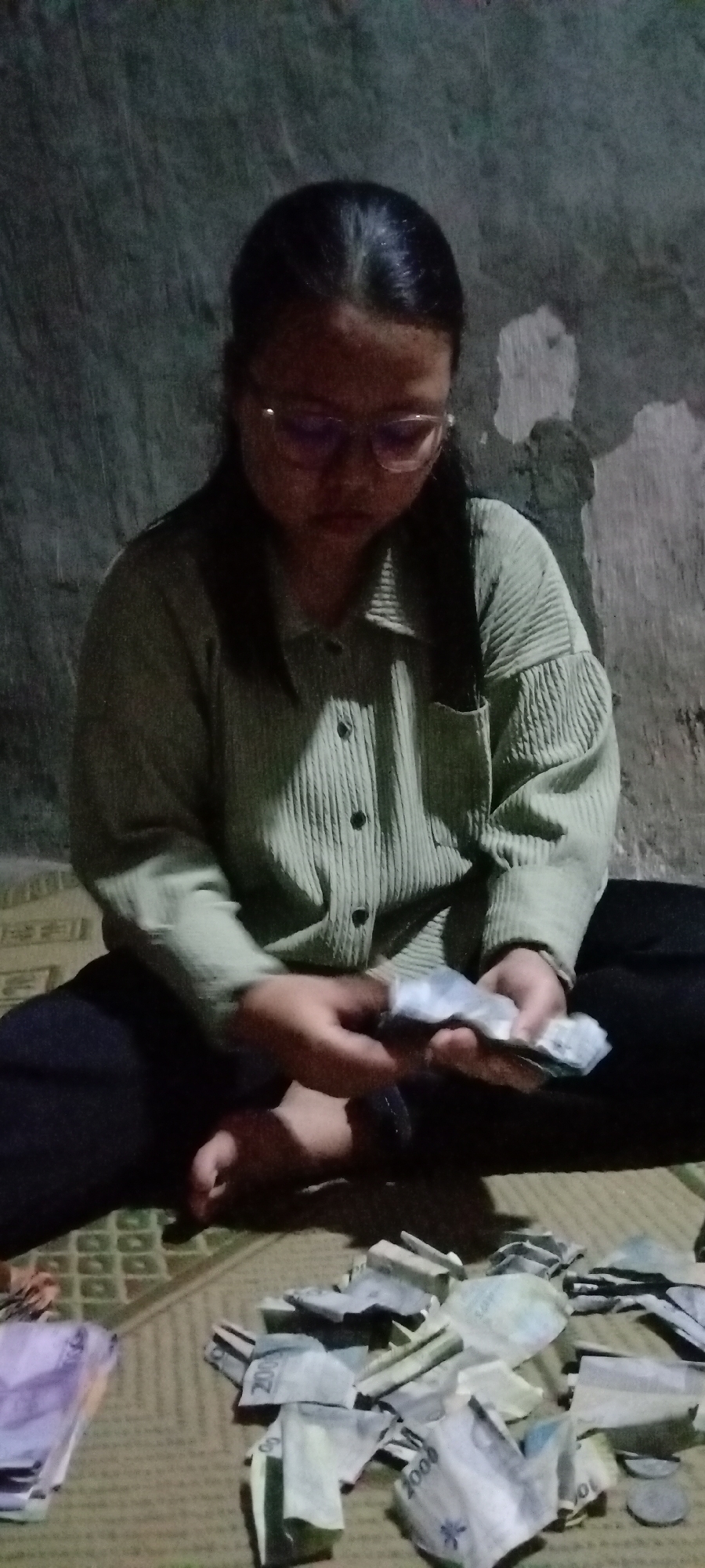 A young woman wearing glasses and a light green corduroy shirt is sitting on a woven mat, counting a pile of Indonesian Rupiah banknotes. She is holding a small stack of bills in her hands, appearing to sort or count them. The background is a dark, textured wall. In front of her, scattered on the mat, are numerous other banknotes, some folded and others appearing to be torn. The lighting is dim, casting shadows on the wall and the woman. The scene suggests a private setting, possibly a home in Utama Jaya, Mataram, Indonesia, where the woman is engaged in managing her finances.