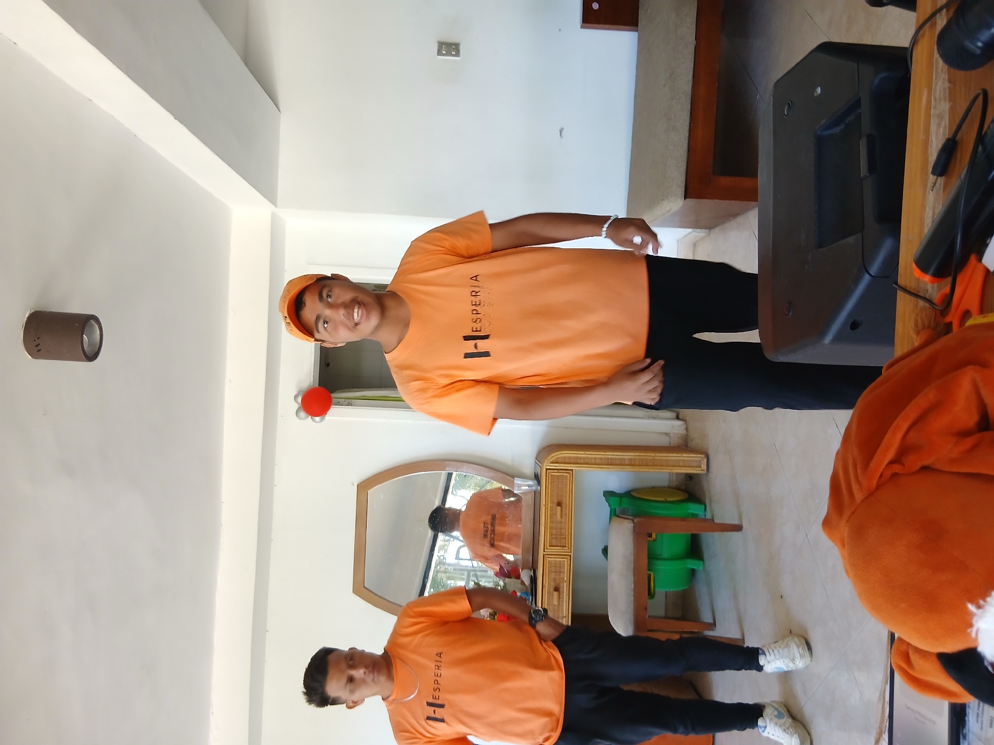 Two young men wearing orange t-shirts with the logo "HESPERIA" stand in a room with white walls and a tiled floor. The man on the left, facing mostly forward with his hands in his pockets, has dark hair and wears black pants and white sneakers. The man on the right, wearing an orange baseball cap and black pants, smiles and faces the camera directly. He has a bracelet on his left wrist. In the background, a dresser with a mirror reflects another person in an orange shirt. A stool with a padded seat sits in front of the dresser. A black speaker is positioned on a wooden table in the foreground. Red and silver balloons are visible on the wall to the right. The scene appears to be indoors, possibly a hotel or event space, in Playa El Agua, Venezuela.