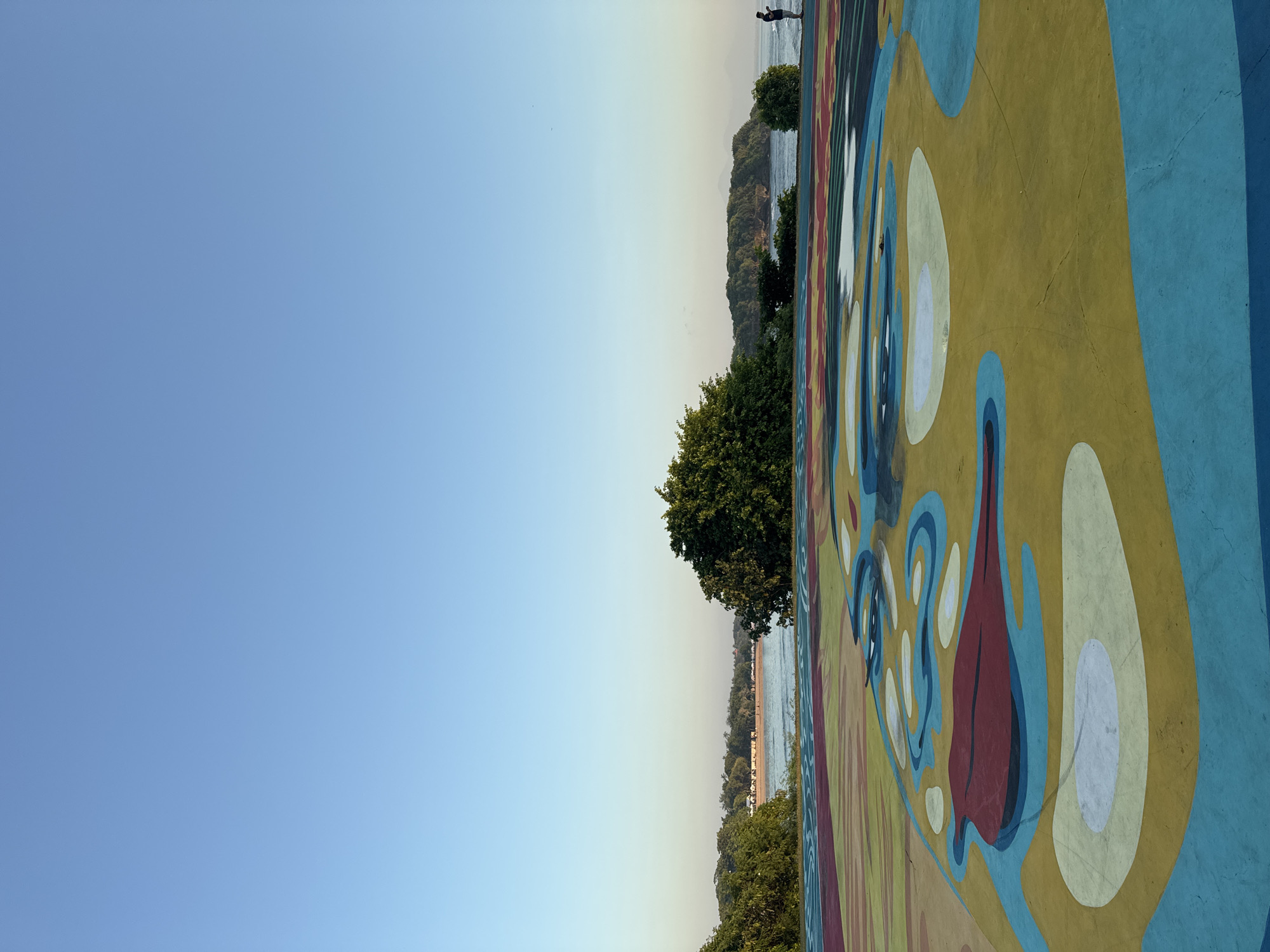 The image shows a mural painting with trees and water in the background. The mural is a vibrant mix of yellow, blue, white, and red colors in abstract shapes. Trees are visible along the horizon line, and water is seen behind the trees. In the background, a figure stands on the edge overlooking the water. The location is Kuta Selatan, Indonesia.