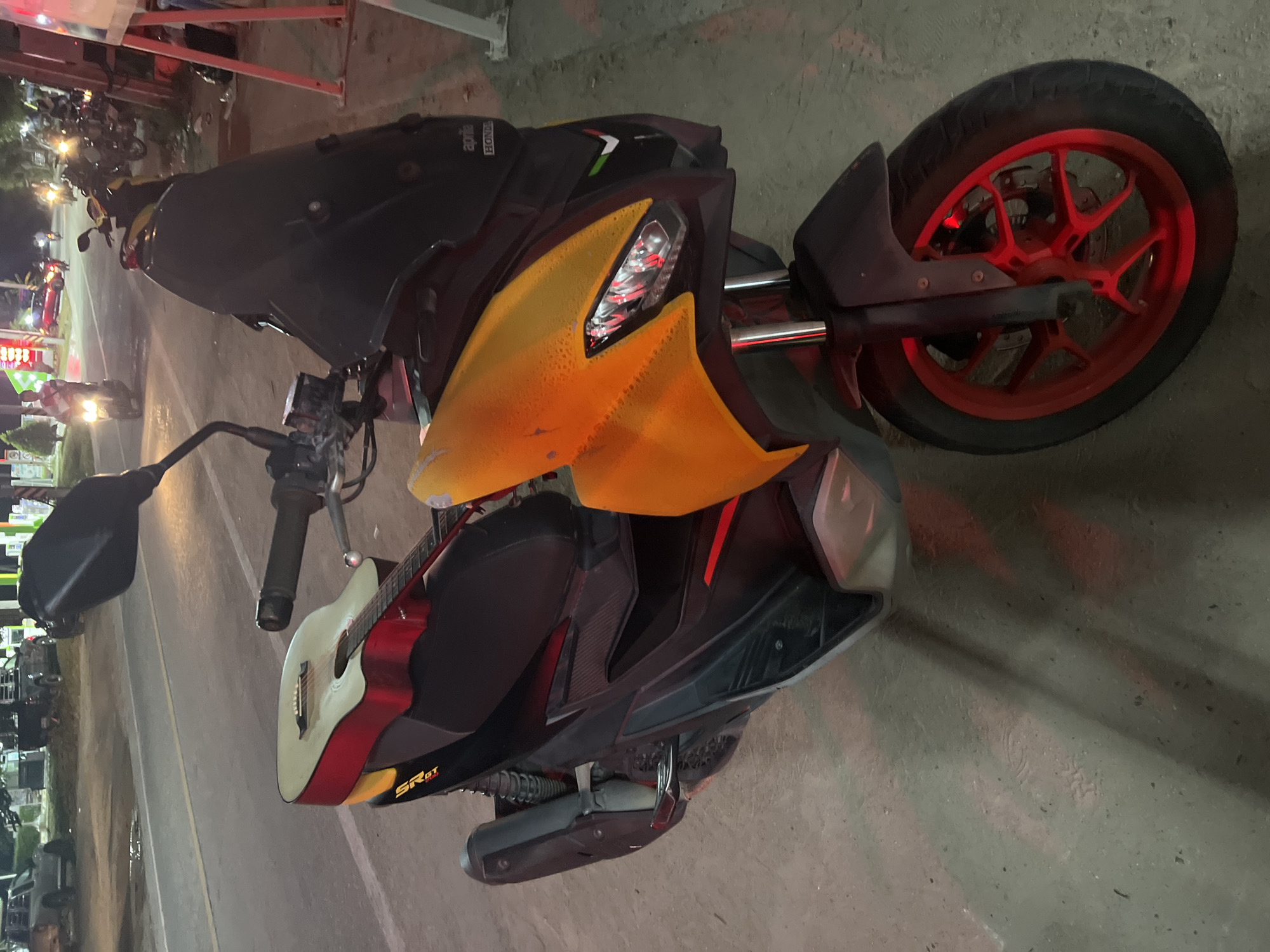 A red and black Aprilia SR GT scooter is parked on a paved surface, possibly a street or parking lot, in Ko Pha-ngan District, Thailand. The scooter has a yellow front fairing and a black windscreen. An acoustic guitar, with a light-colored top and red sides, is resting on the seat of the scooter. The scooter's front wheel has a red rim, and the scooter is illuminated by red light, likely from nearby signage or streetlights. In the background, other vehicles and a roadside establishment are visible. The overall scene suggests an urban or semi-urban environment at night.