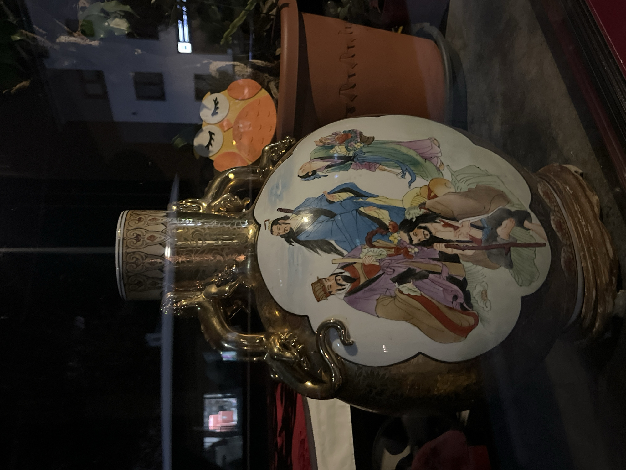 The image shows a decorative vase with a painted scene of figures in traditional Chinese attire. The vase has golden handles that resemble dragons or serpents. The scene on the vase depicts several men and women, some holding objects like staffs and baskets of flowers. The vase is positioned in what appears to be a display window, with reflections of a nighttime urban environment visible. To the right of the vase, a terracotta pot containing a plant is visible, and behind that, a building with windows and lights can be seen. An orange owl decoration is placed in front of the plant. The scene is illuminated by artificial light, suggesting it is evening or nighttime. The setting is a shop window in the city of Saint-Vith, Belgium.