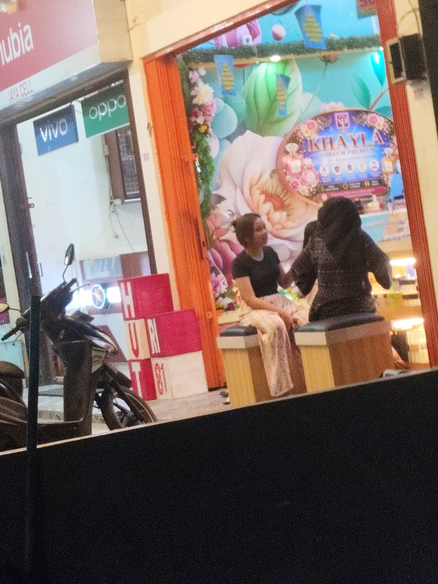 A slightly blurry, low-light image shows the exterior of a shop in Palembang, Indonesia. The shop appears to be a retail store selling mobile phones and possibly perfumes. To the left, a sign reads "JAYA CELL" with "nubis" above it. Below this are logos for "VIVO" and "OPPO". A motorcycle is parked in front of the shop. To the right, the shop entrance is adorned with orange curtains and a large floral mural featuring the brand name "KHAYLA PARFUM PREMIUM". Two individuals are visible inside the shop; one, a woman in a black t-shirt and patterned pants, is seated, facing a second individual who is seated and facing away from the camera. The second individual is wearing a dark, hooded garment. Behind them, shelves with products are illuminated by display lights. Red and white stacked boxes with "HURT 81" printed on them are visible to the left of the entrance. The overall scene suggests a commercial establishment on a street in Palembang.
