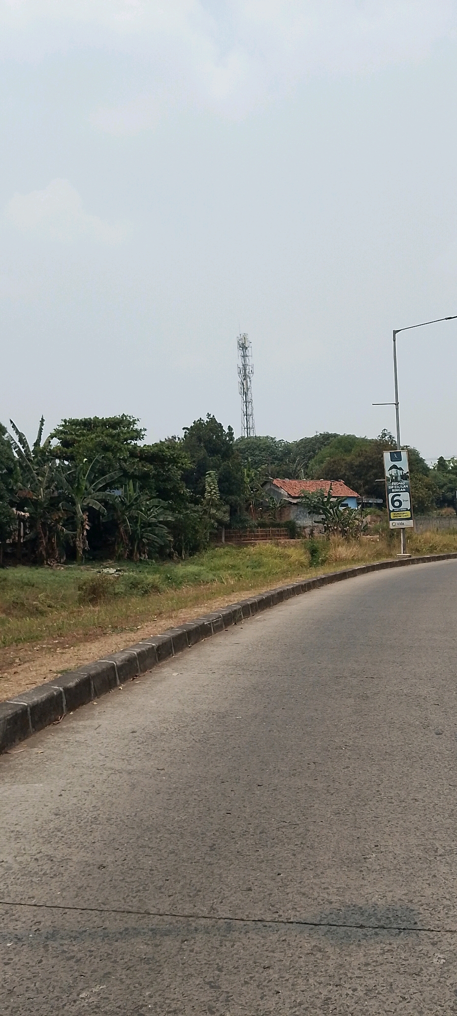 The image shows a roadside scene in Bekasi, Indonesia. A paved road curves to the right, bordered by a concrete curb and a grassy area. In the background, there is lush green vegetation, including banana trees, and a tall telecommunications tower. A small house with a red roof is nestled amongst the trees. A street light with a sign is visible on the right side of the road. The sky is overcast.