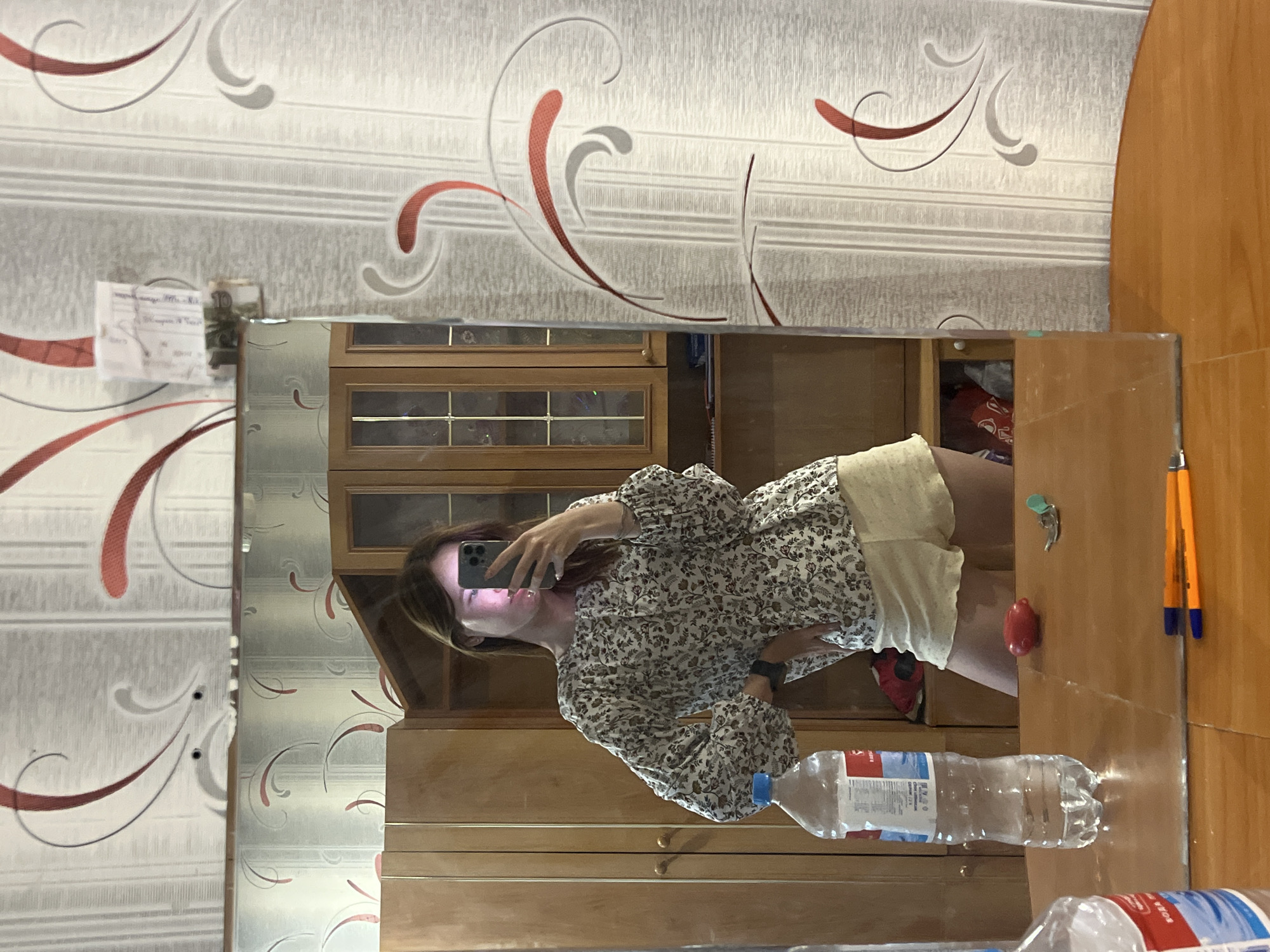 A young woman is taking a selfie in front of a mirrored wardrobe. The mirror reflects an interior scene with patterned wallpaper. The wardrobe has a wooden frame with cabinets. On top of the wardrobe are pens, water bottles, and a red object. The location is Chaikovsky, Russia.